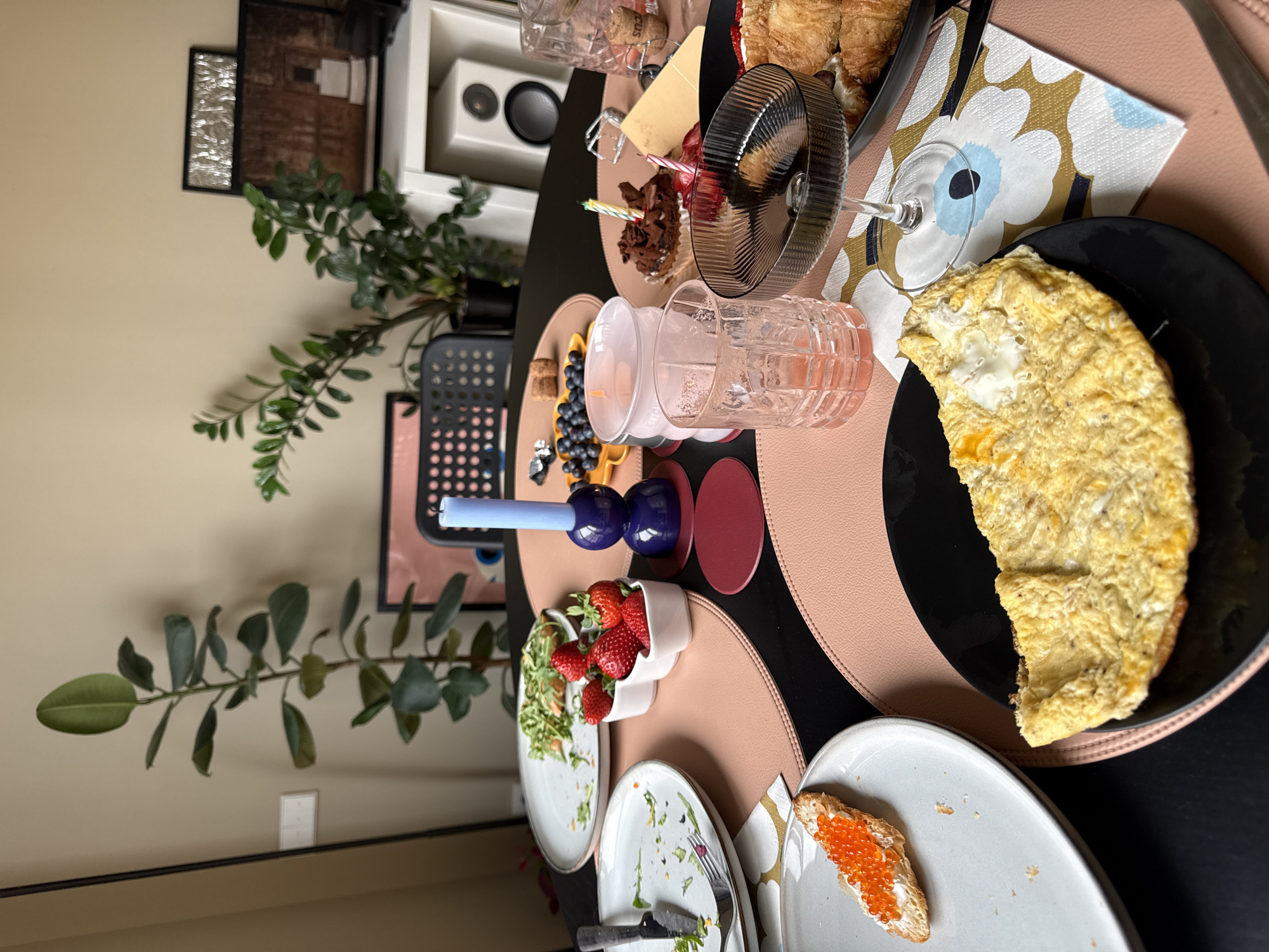 The image depicts a table setting with various food items. An omelet sits on a black plate, next to a slice of bread topped with orange roe. There is also a croissant, a dessert with lit candles, and a bowl of strawberries, along with blueberries and other small pastries. Empty plates with remnants of a meal are visible. Several glasses and a blue decorative candle holder are also on the table.
In the background, there is a potted plant, a black chair, artwork, and a white speaker. The setting suggests a meal in a domestic environment.
There is no clear indication of the location being Barcelona, Spain.