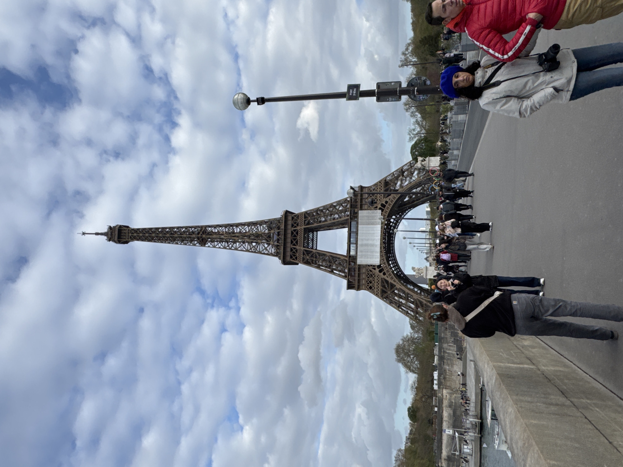 The image shows the Eiffel Tower in Paris, France, from a bridge. Several people are on the bridge, including a couple posing for a photo; the woman is wearing a blue beret. Some people are taking pictures. The sky is cloudy. Street lights are on the bridge.