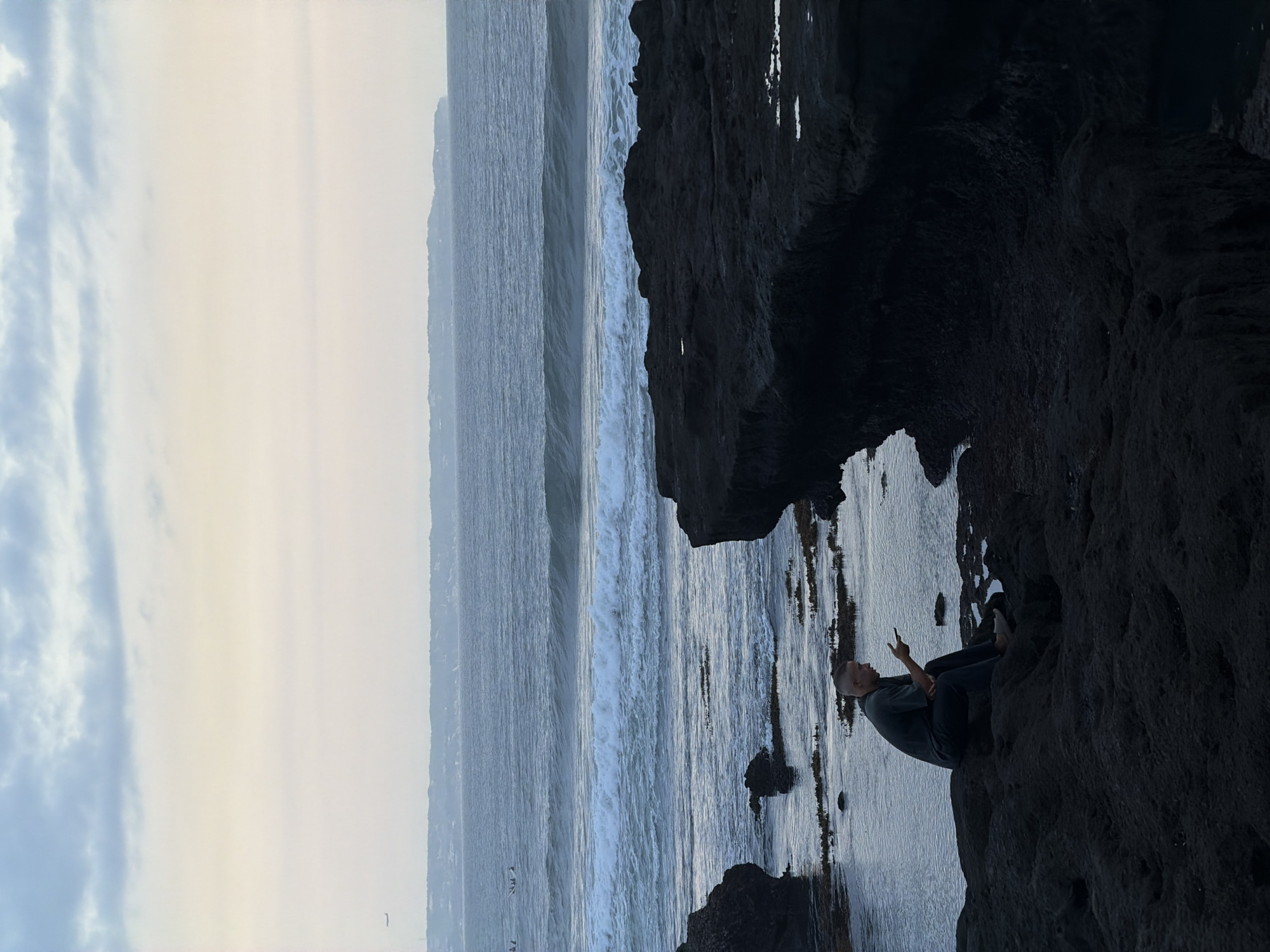 The image shows a person sitting on a dark rocky outcrop next to a beach in North Kuta, Indonesia. The sky is overcast with a light haze. The ocean has moderate waves. The person is facing the ocean and holding up two fingers in a "peace" sign. The tide appears to be low, with some rocks exposed.