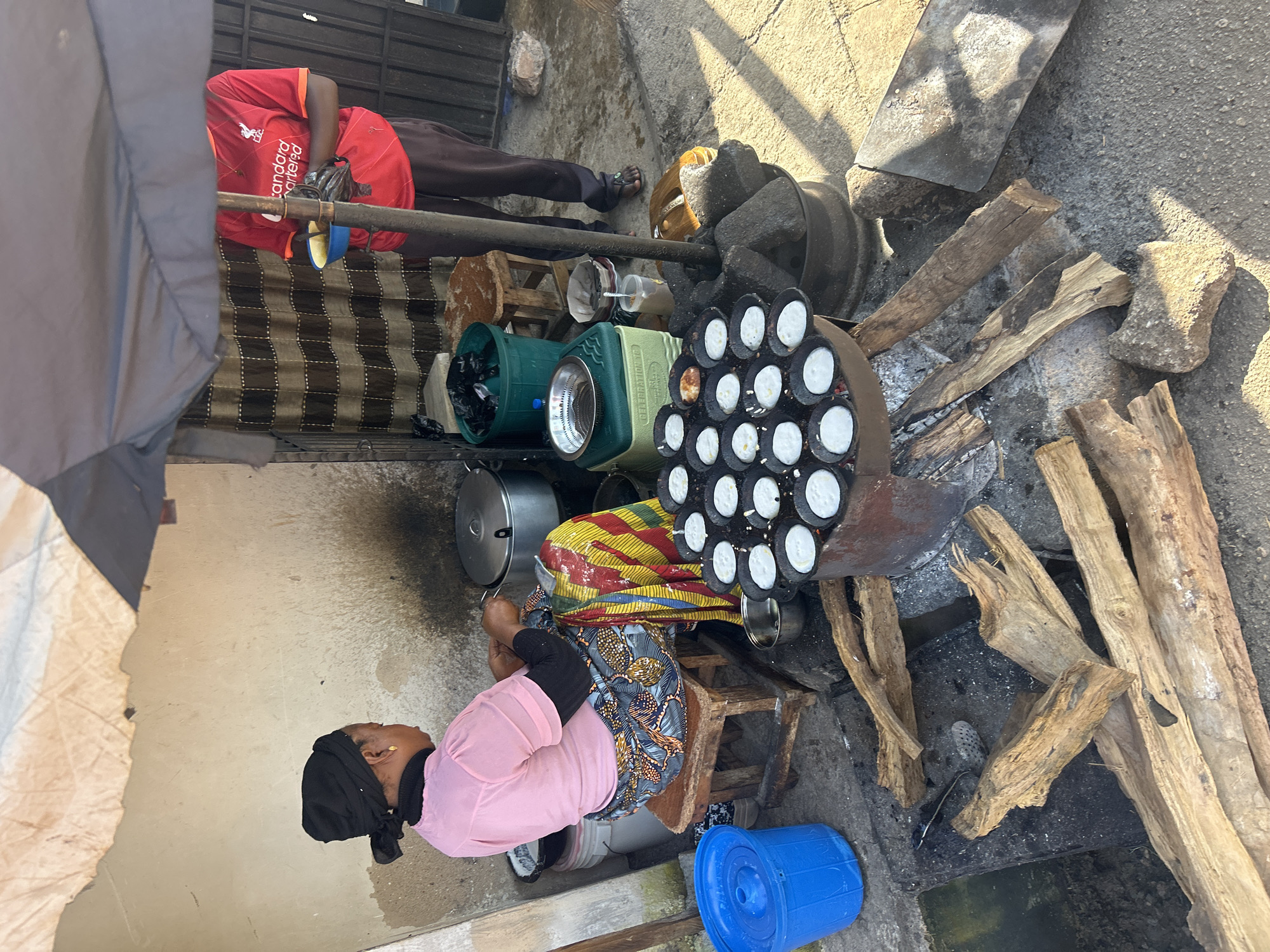 This image depicts an outdoor food stall in Jos, Nigeria. A woman wearing a black headscarf and a pink shirt sits on a wooden stool, preparing food in a circular pan over an open fire. She is wearing a colorful patterned skirt. To her right, another person in a red Liverpool FC jersey stands under an umbrella, holding a small bowl. Various cooking utensils, pots, and containers are visible around the stall, including a large blue bucket. The ground is concrete, and there are piles of firewood near the cooking area. The overall scene suggests a bustling street food environment.