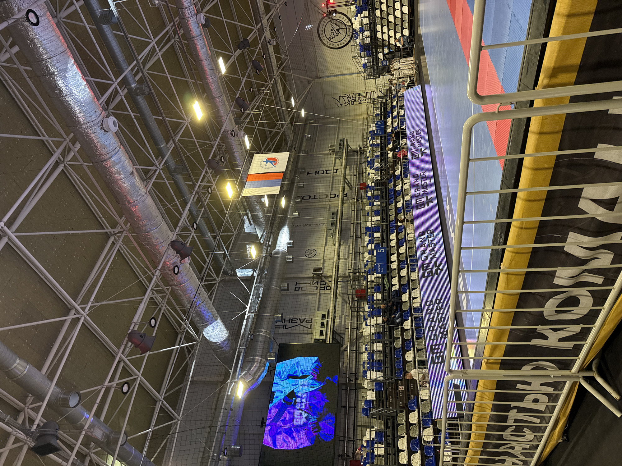 The image shows an indoor arena, likely for sports or events, in Rostov-on-Don, Russia. Overhead, there is a complex network of metal beams, pipes, and lights. A Russian flag hangs from the ceiling. There is a large screen displaying graphics or video, and stadium-style seating is visible, some of which is occupied. A sign with "GRAND MASTER" and Russian words is displayed vertically. A metal railing is in the foreground.