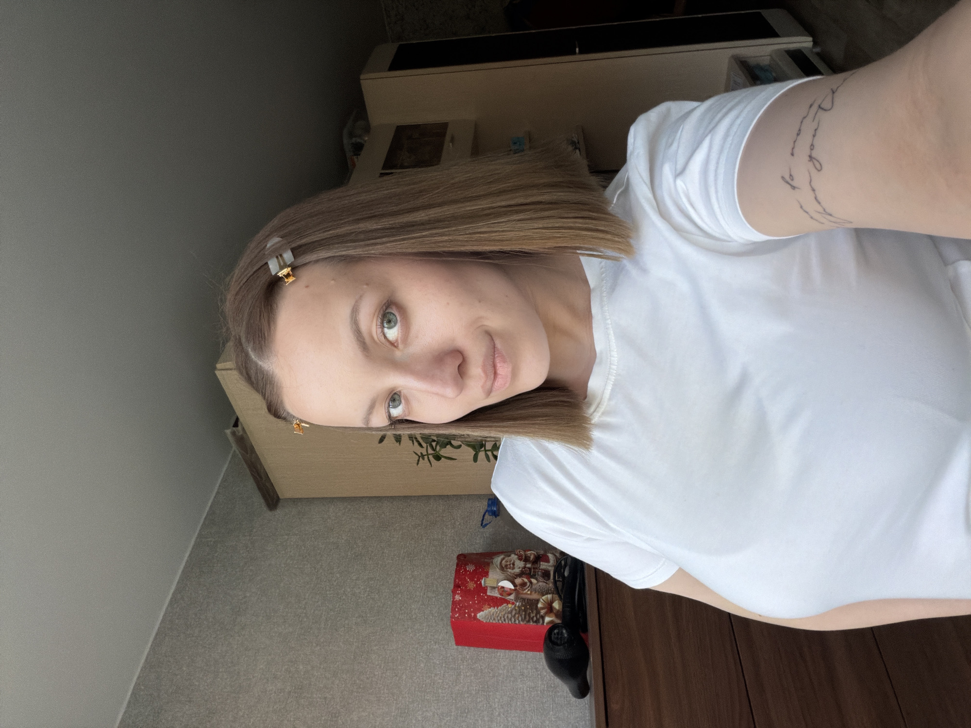 A woman is posing for a selfie indoors. She has short, light brown hair with gold-colored hair clips on the left side of her head. She is wearing a white t-shirt. There is a tattoo on her upper right arm.
Behind her, various items are visible including furniture and a red box with a Christmas-themed design. The background is somewhat blurred. It is not possible to verify the location or the city of Раздолье, Russia.
