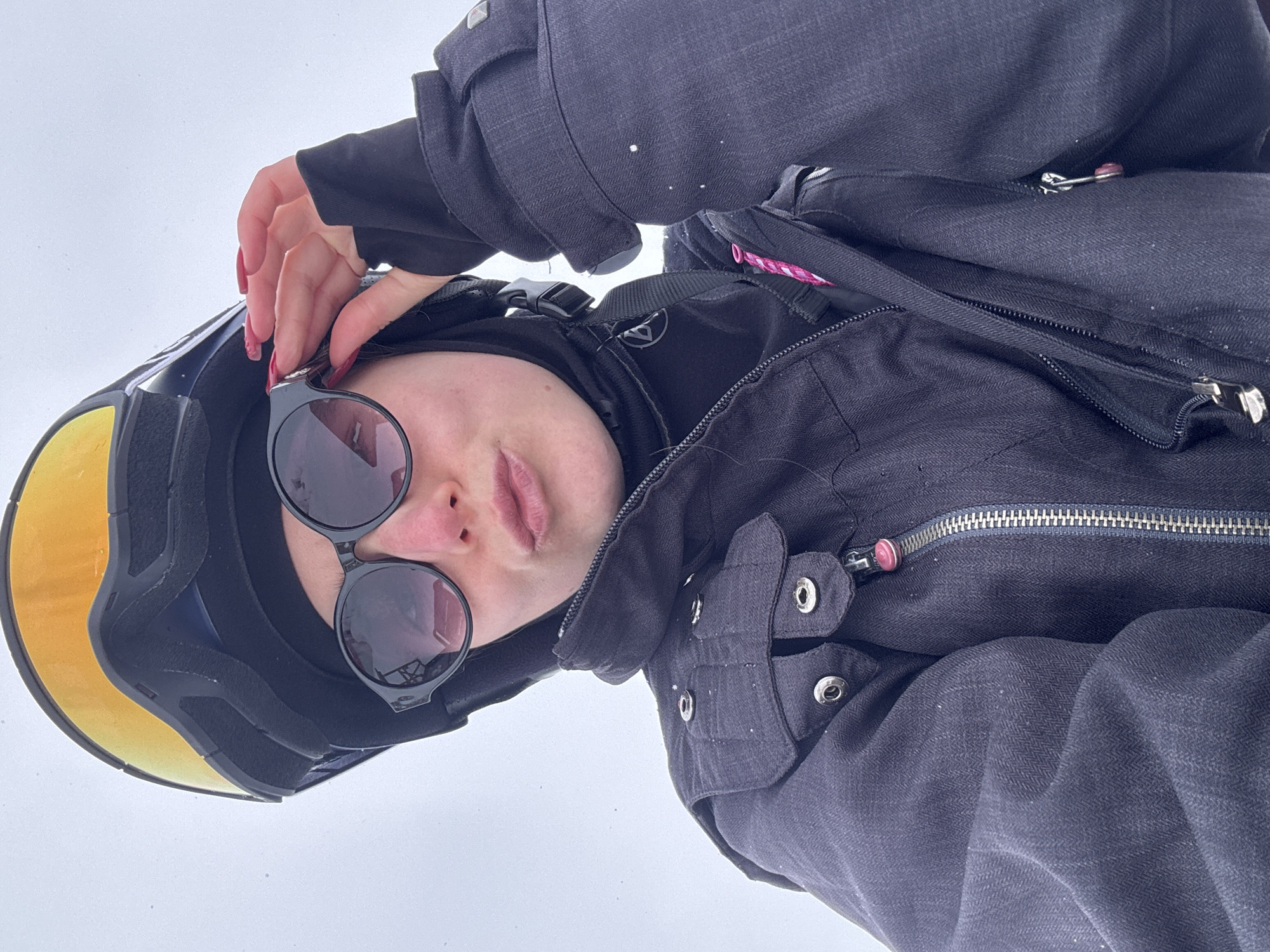 A person is looking down at the camera while standing in a snowy environment. The individual is wearing ski goggles with a yellow lens, a black head covering, and round-frame sunglasses. They are also dressed in a dark jacket with visible zippers, buttons, and a backpack strap. The backdrop is a plain, white expanse suggesting a snow-covered landscape. The location is Unknown, Unknown.