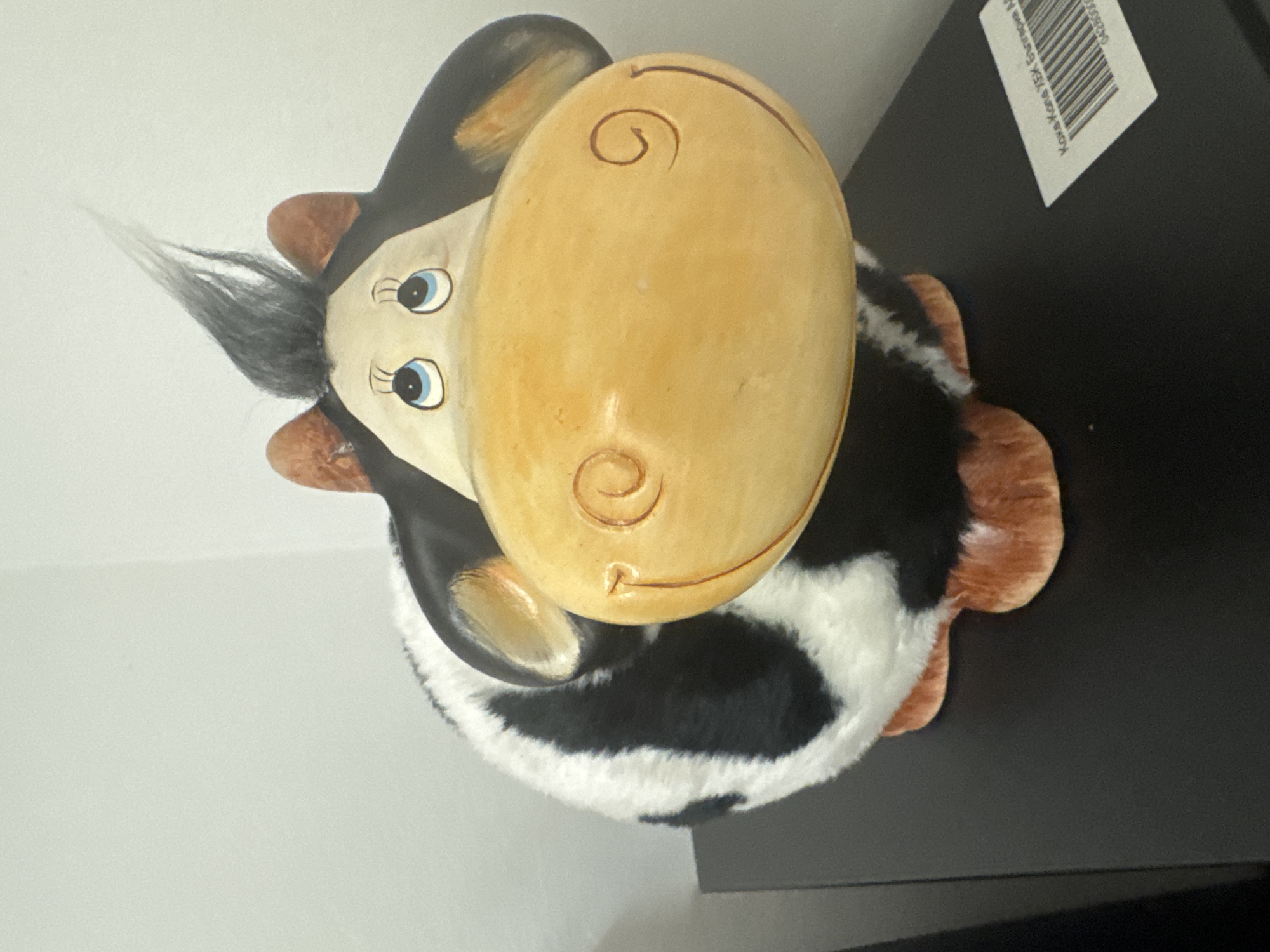 A cow figurine is on a dark surface. The cow has a large, light yellow nose, black and white fur, brown ears, and blue eyes with eyelashes. It also has tufts of grey hair sticking up from its head. The background is a light gray wall on one side and a black surface on the other, with a white sticker containing a barcode visible in the upper right corner of the frame. The figurine appears to be located indoors, possibly on a shelf or table. It is difficult to ascertain the exact location in Sofia, Bulgaria, based solely on the provided image.