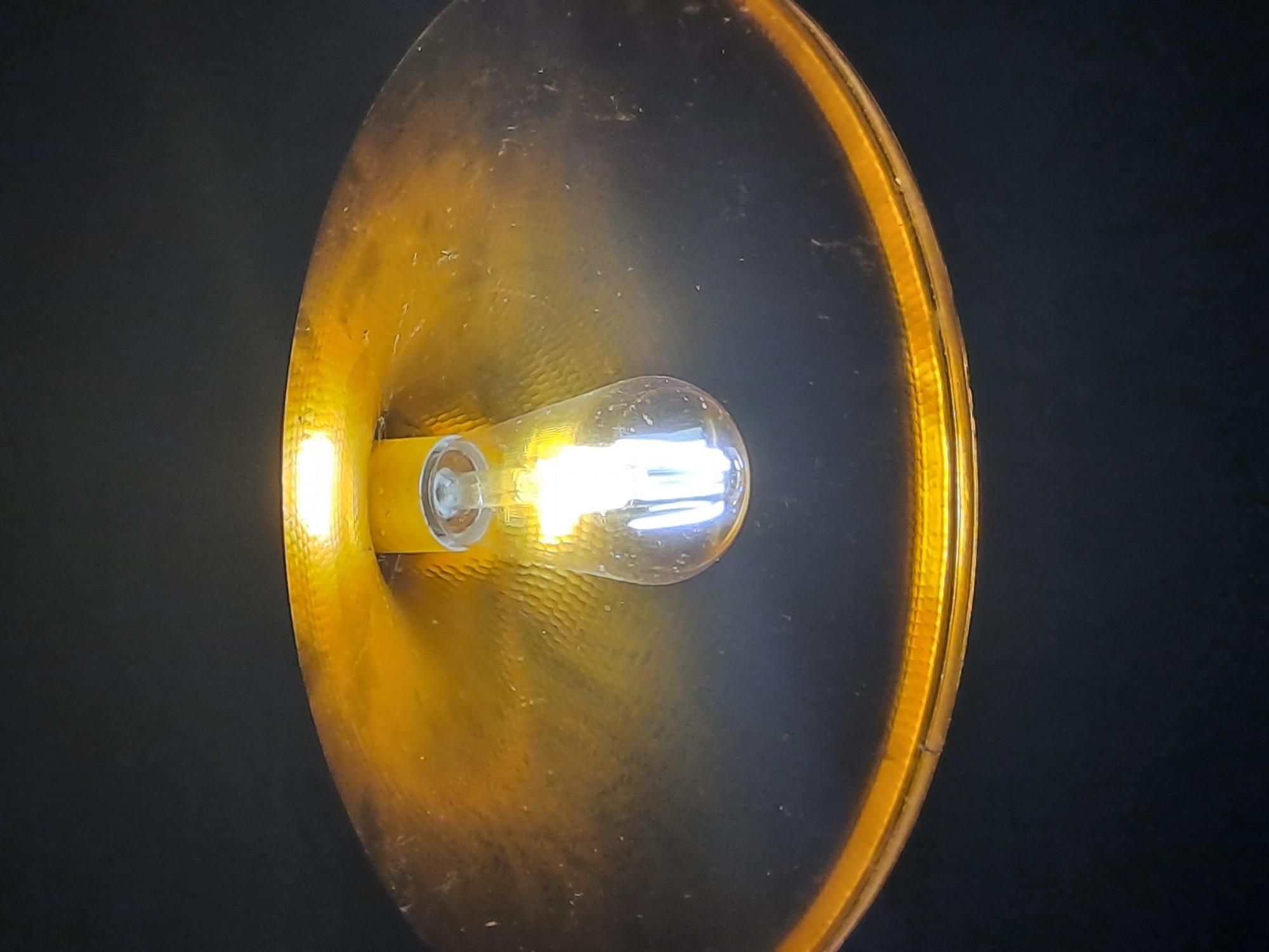 A close-up, top-down view of a single light fixture with a metallic gold shade hangs against a dark background, likely the ceiling of a room in Krasnyi Ogorok, Russia. A clear, incandescent light bulb illuminates the interior of the shade, casting a warm glow. The shade itself has a hammered texture, and dust particles are visible on its surface. The light bulb is suspended by a white fixture. The scene appears to be an indoor domestic setting.