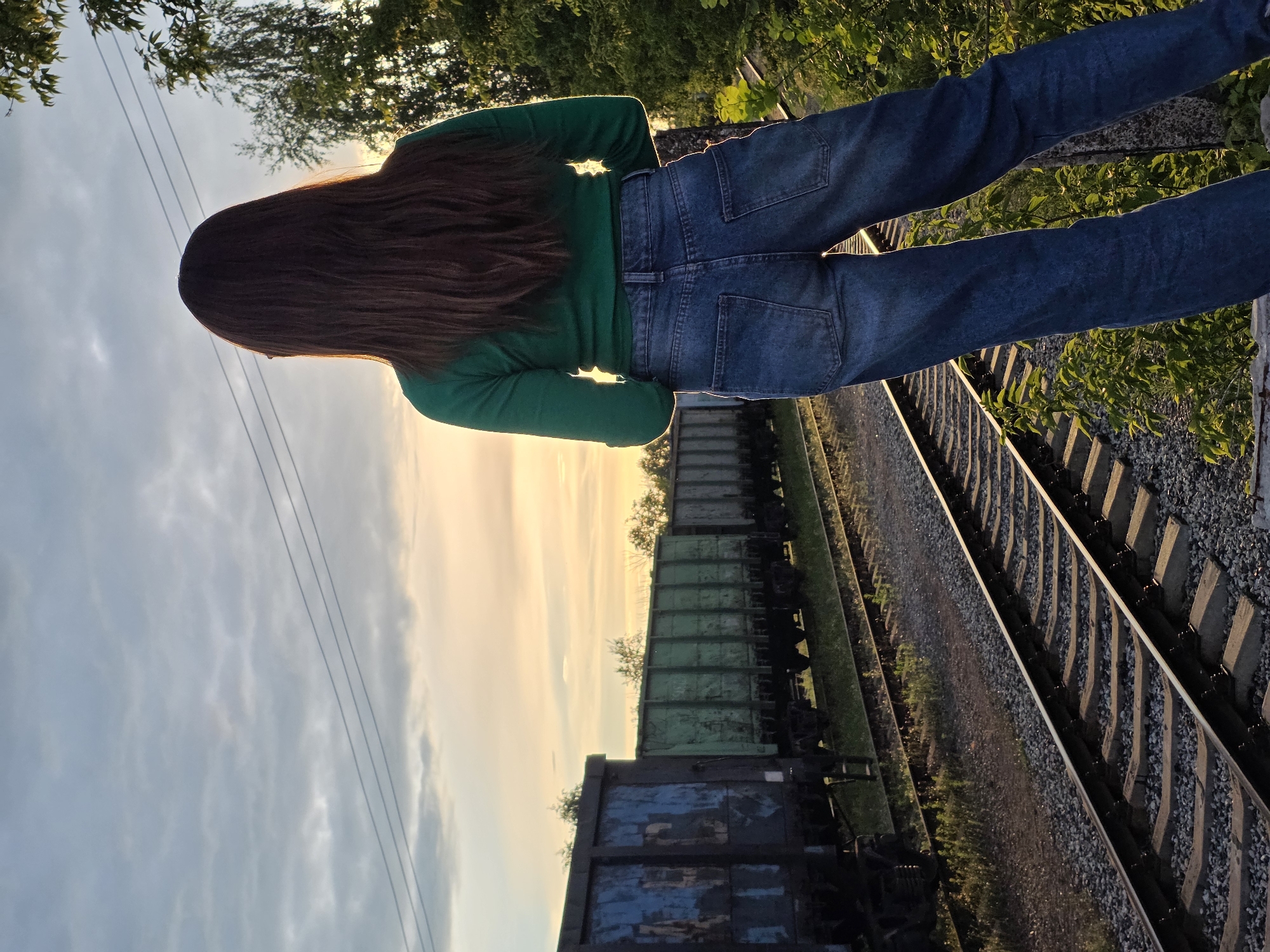 This is a vertical, outdoor, eye-level, full-shot photograph of a person standing on ballast next to a railway track, with their back to the camera. The person is wearing a green long-sleeved shirt and blue denim jeans. Their long, dark brown hair cascades down their back. To the left of the frame, a vast sky with scattered clouds stretches out, illuminated by the warm glow of sunset or sunrise. Three power lines are visible against the sky. To the right of the person, several freight train cars are parked on parallel tracks. The closest car is dark blue with peeling blue paint. The next car is a weathered green hopper car. The tracks themselves are lined with concrete ties and covered in gravel, with a strip of green grass bordering one side. Trees and foliage are visible in the background, particularly to the upper right of the frame. The lighting suggests it is either early morning or late evening. The overall mood is serene and contemplative. No text is visible in the image.