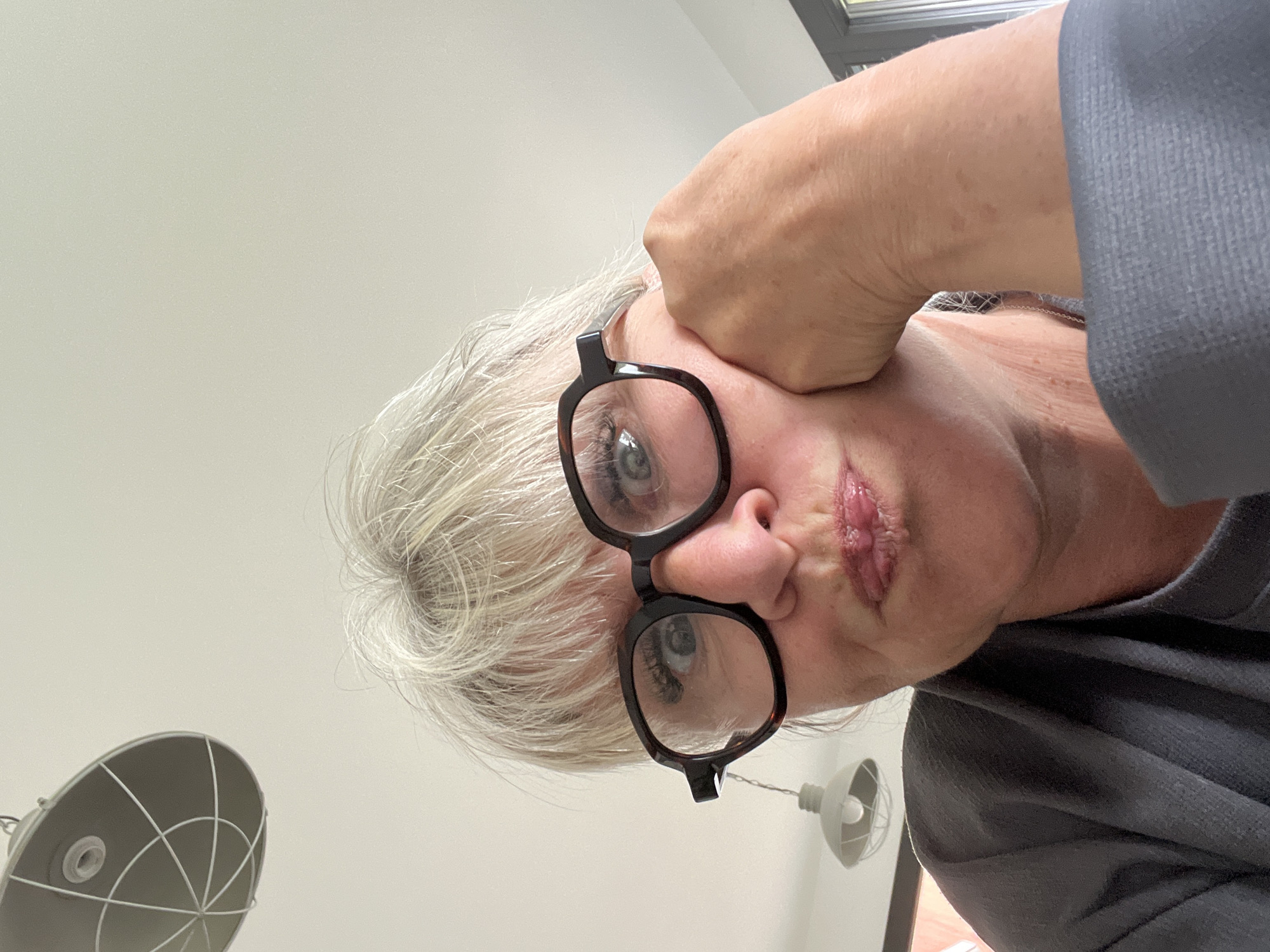 A close-up, low-angle, indoor photograph depicts a person with light skin, gray hair, and black-framed glasses. Their left hand is positioned against their forehead, near the hairline. The person's mouth is slightly pursed. They wear a gray garment. In the background, parts of two light fixtures are visible against a light-colored ceiling. The photo's location, Chambœuf, France, cannot be verified from the visual information.