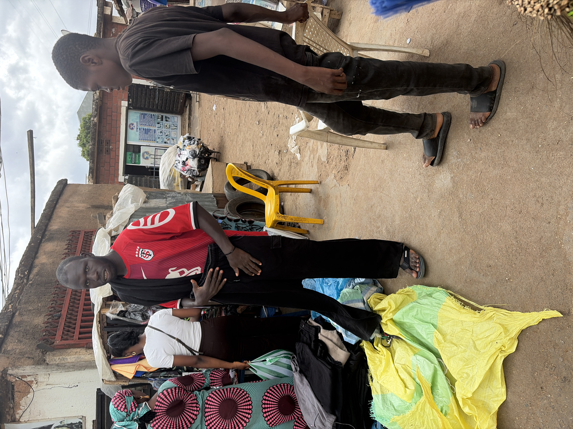 The photograph captures an outdoor market scene in Jos, Nigeria, on an overcast day. The ground is unpaved and dusty, suggesting an informal commercial area.
Two men are central to the image. On the left, a young man with short dark hair wears a dark grey t-shirt and black pants, with black slip-on sandals. He stands slightly hunched, looking downwards. On the right, another man, also with short dark hair and a neutral expression, wears a red short-sleeved soccer jersey with white and black accents. The jersey features a Nike swoosh, what appears to be the Toulouse FC club crest (a red shield with a white emblem), and the text "SYNERGY" below it, with the number "8" visible on the sleeve. He is wearing black pants and a single black flip-flop on his visible foot. He holds up a pair of black pants, seemingly displaying them for sale.
Partially visible behind the man in the red jersey is a woman wearing a white t-shirt and dark skirt, with her back mostly to the camera. In the lower right corner, another person is seen, clad in a vibrant blue, pink, and white patterned garment.
Around the individuals, various goods are displayed. Piles of clothing, some wrapped in plastic bags, are scattered on the ground. A large, crinkled yellow and light green tarp or bag lies prominently in the foreground. Several old tires are visible near a yellow plastic chair. In the background, dilapidated buildings with brick walls and tin roofs line the street. A sign on one building's wall displays text and several small pictorial icons, though the text is illegible due to distance. Overhead wires crisscross the sky. A white plastic chair is also seen in the mid-ground.