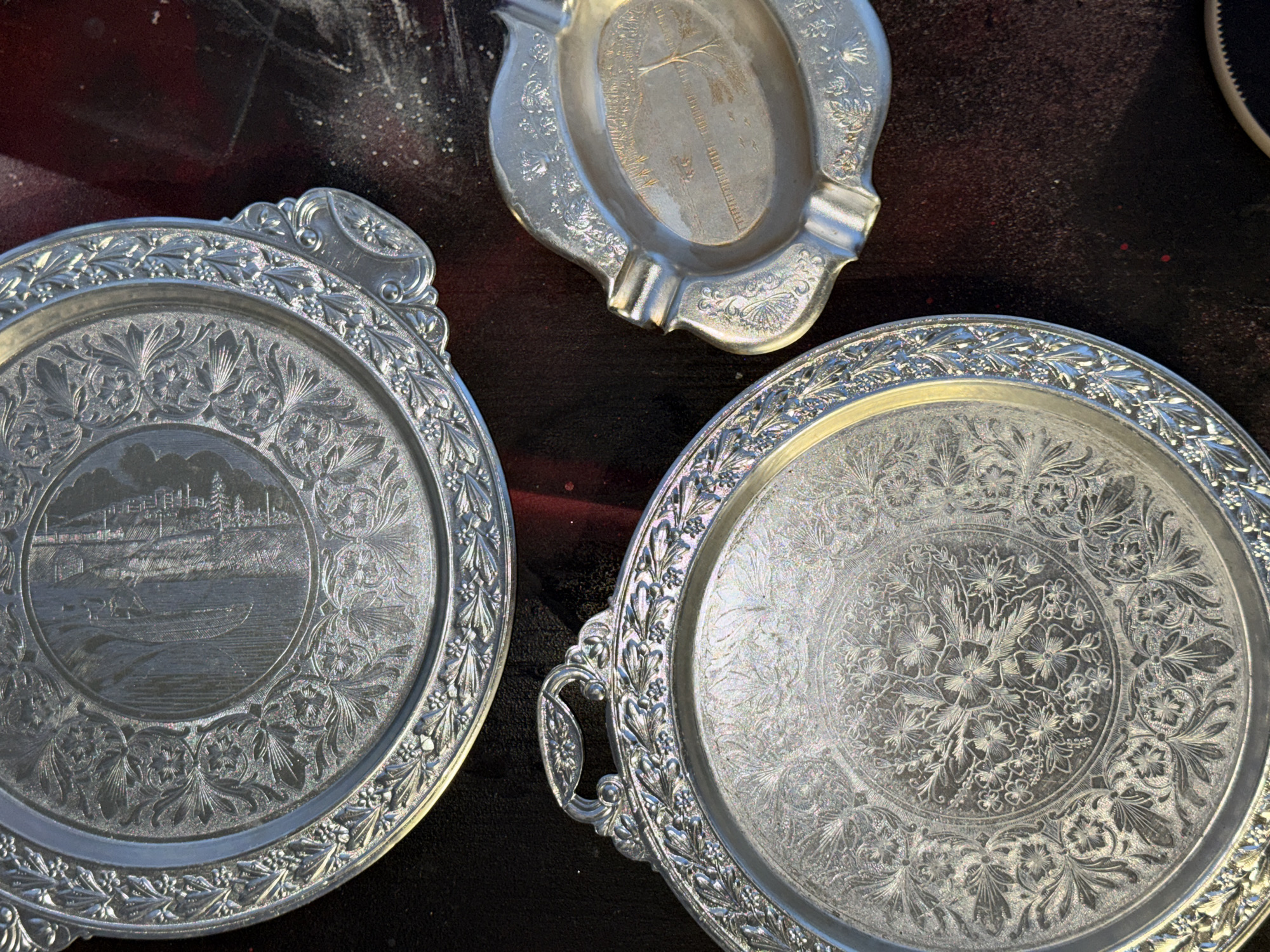 The image shows three tarnished silver trays on a dark, slightly speckled surface. The trays are ornate, featuring raised designs and intricate detailing. One tray is round with two handles and a floral design in its center. Another is also round but has a detailed cityscape scene in its center, possibly depicting a body of water with buildings in the background. The third tray is smaller, oval-shaped, and has a worn-out tree image in its center. Based on the buildings depicted on one of the trays, the location is likely Krasnoyarsk, Russia.