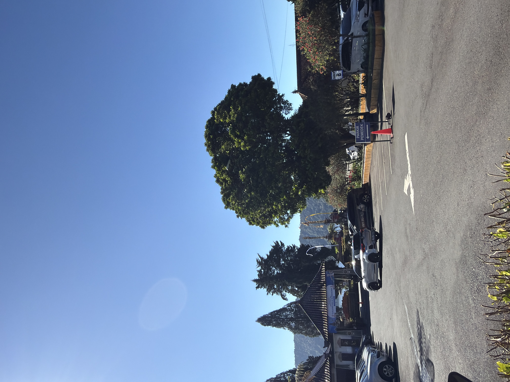 The image shows a parking lot in Baturiti, Indonesia, on a clear day. The sky is bright blue with a slight lens flare. In the foreground, there is an asphalt parking lot with white lane markings and a few cars. A white SUV is seen driving away from the camera. In the middle ground, there are several trees, including a large, leafy tree on the right and a tall, conical tree on the left. There are also some buildings with traditional roofs. A sign that reads "PARKIR PEMEDEK" with an arrow pointing to the left is visible. In the background, there are mountains covered in lush vegetation. The scene appears to be a tourist area, possibly a temple or a scenic viewpoint, given the traditional architecture and the presence of visitors' vehicles.
