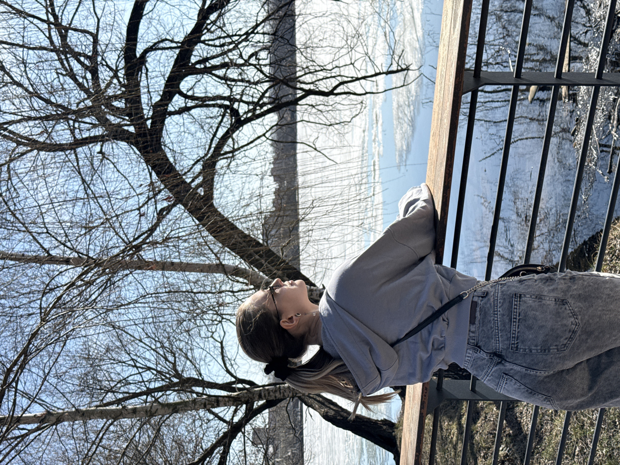 A young woman stands facing away from the camera, leaning against a wooden railing overlooking a body of water and bare trees against a light blue sky. She wears a light gray sweatshirt, blue jeans, and glasses, with her hair pulled back in a ponytail. A black crossbody bag hangs at her side. The background shows what appears to be a partially frozen body of water and leafless trees. The presence of a metal railing suggests she is on a bridge or observation platform. The location is the embankment of the Moskva River in the city of Moscow, Russia.