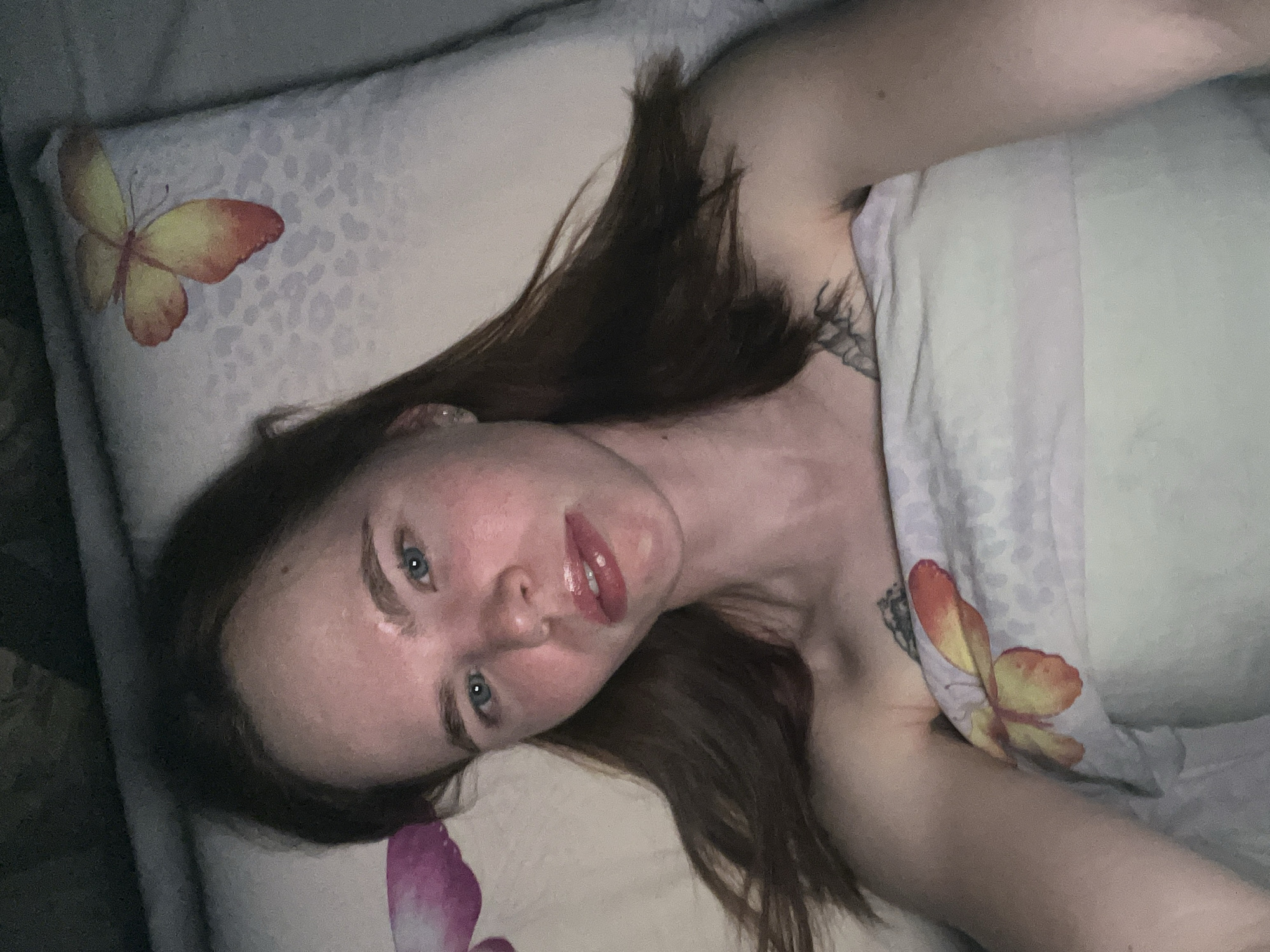 A young woman lies in bed, resting her head on a pillow adorned with butterfly prints. She is partially covered by a light-colored blanket, also decorated with a butterfly design. Her hair is brown, and she has fair skin and blue eyes. The photograph seems to be taken indoors, likely in a residential setting. The location is unknown.