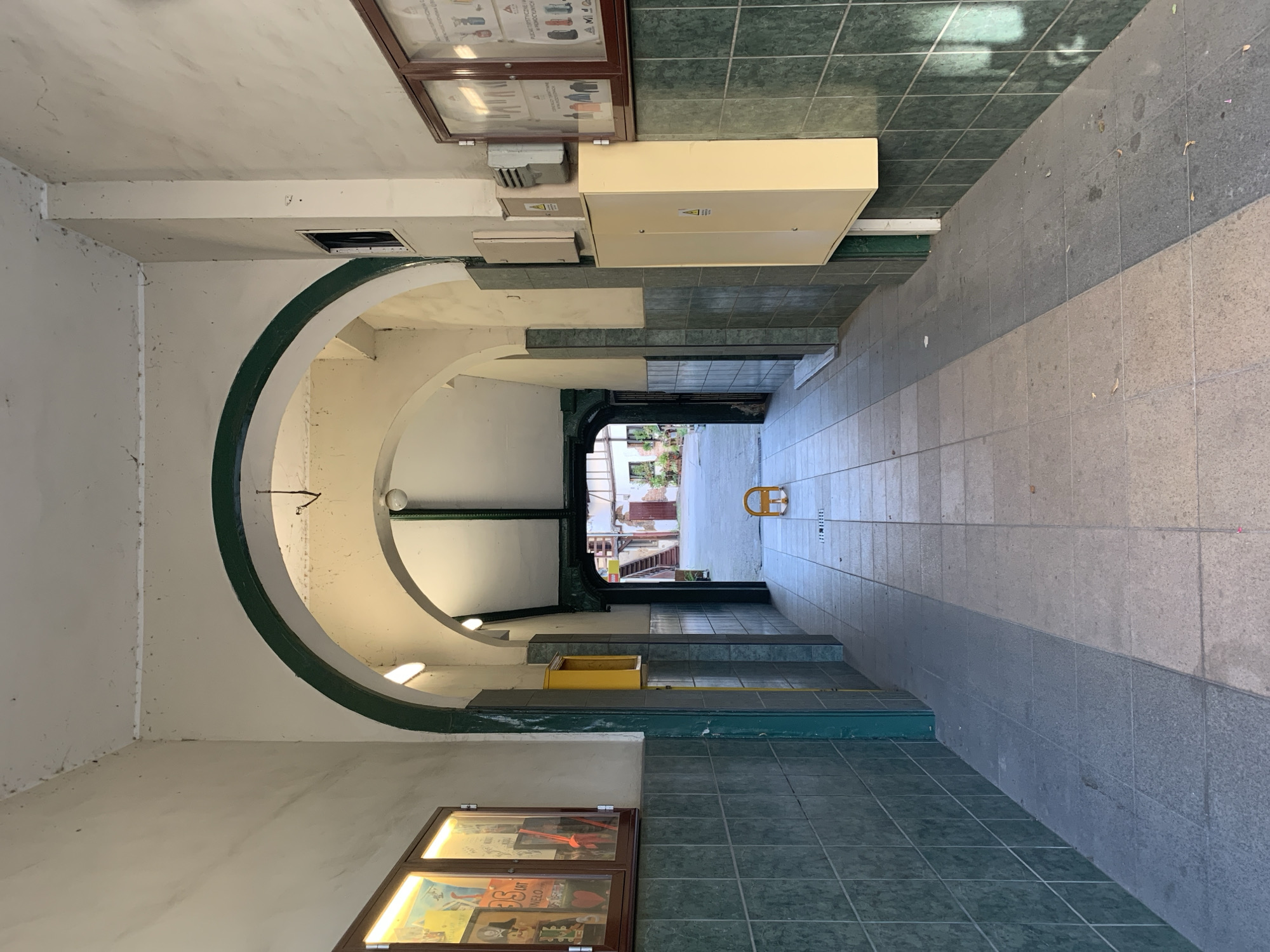 The image depicts a covered walkway or corridor with arched ceilings. The walls are tiled with dark green square tiles up to a certain height, and the upper portions are painted a light, off-white color, showing signs of age and some staining. The ceiling is also off-white and features some visible electrical conduits and lighting fixtures. Green trim accents the arches and vertical structural elements.
On the left wall, there is a framed display case illuminated from within, containing various colorful posters or artwork. Further down the corridor, a bright yellow rectangular box is mounted on the wall. On the right wall, there are several electrical boxes, including a larger beige one and smaller grey and white units. Another framed display case is mounted on the right wall, containing what appear to be informational charts or diagrams with illustrations.
The floor of the corridor is paved with rectangular tiles, arranged in a pattern. The lighting suggests it is daytime, with sunlight casting distinct shadows on the floor, indicating an opening at the far end. In the middle of the corridor, near the far opening, a small, bright yellow parking barrier is present.
The walkway leads to an open courtyard or exterior area where more buildings, stairs, and doorways are visible. The overall impression is of an older building's internal or connecting passageway. The presence of a parking barrier suggests this might be an area with access for vehicles or designated parking.
This covered walkway is located at the courtyard of the building at the address Poznańska 7 in Poznań, Poland. The building is known for its historical significance and is part of the urban fabric of the city.