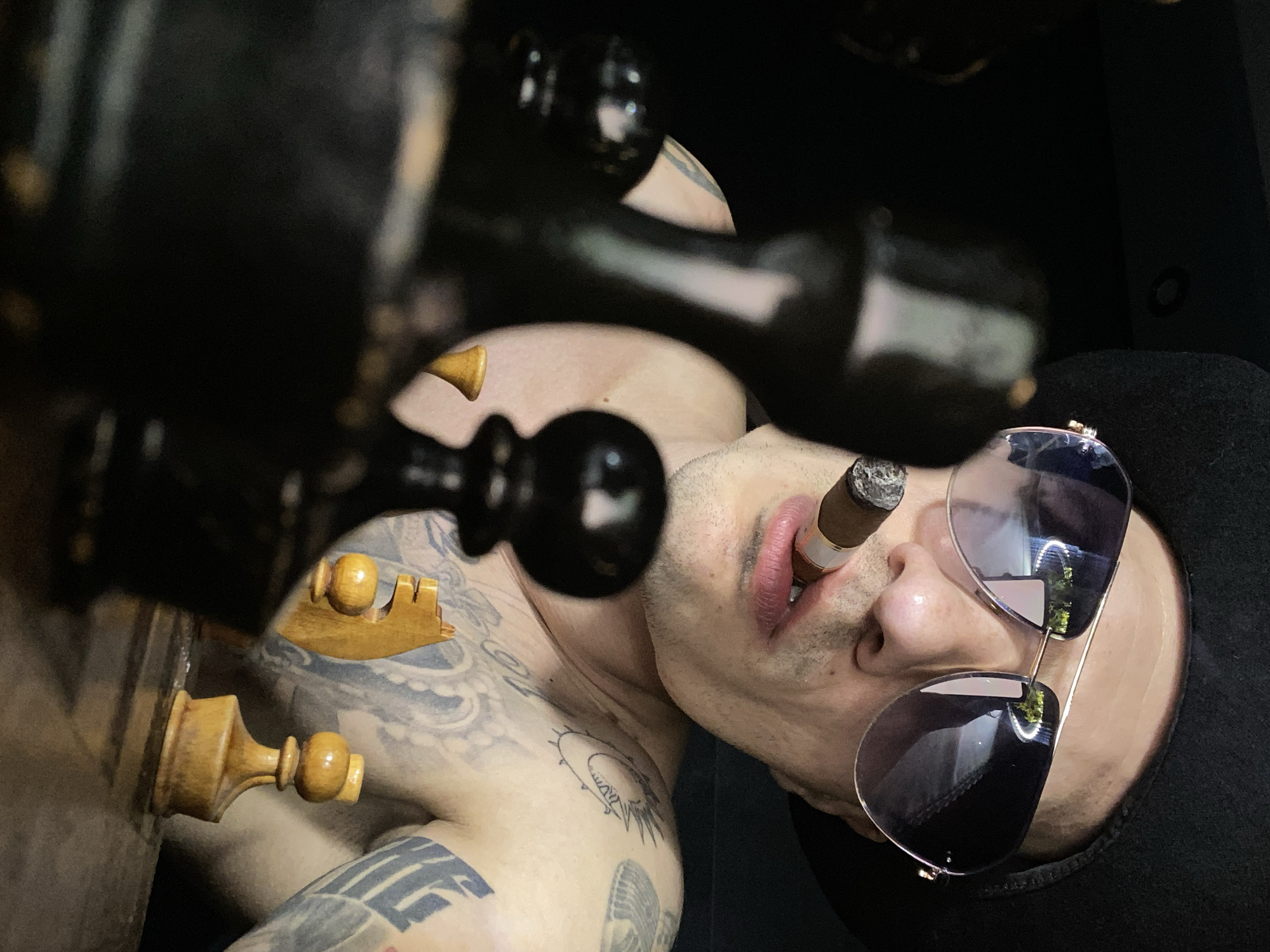A low-angle close-up shot depicts a shirtless, light-skinned individual with visible tattoos, a cigar in their mouth, and sunglasses resting on their face. Several chess pieces, both black and white, are positioned prominently in the foreground and over the individual's body. The background appears to be a dark, solid color. The location is not discernible as Pleven, Bulgaria, from the given image.