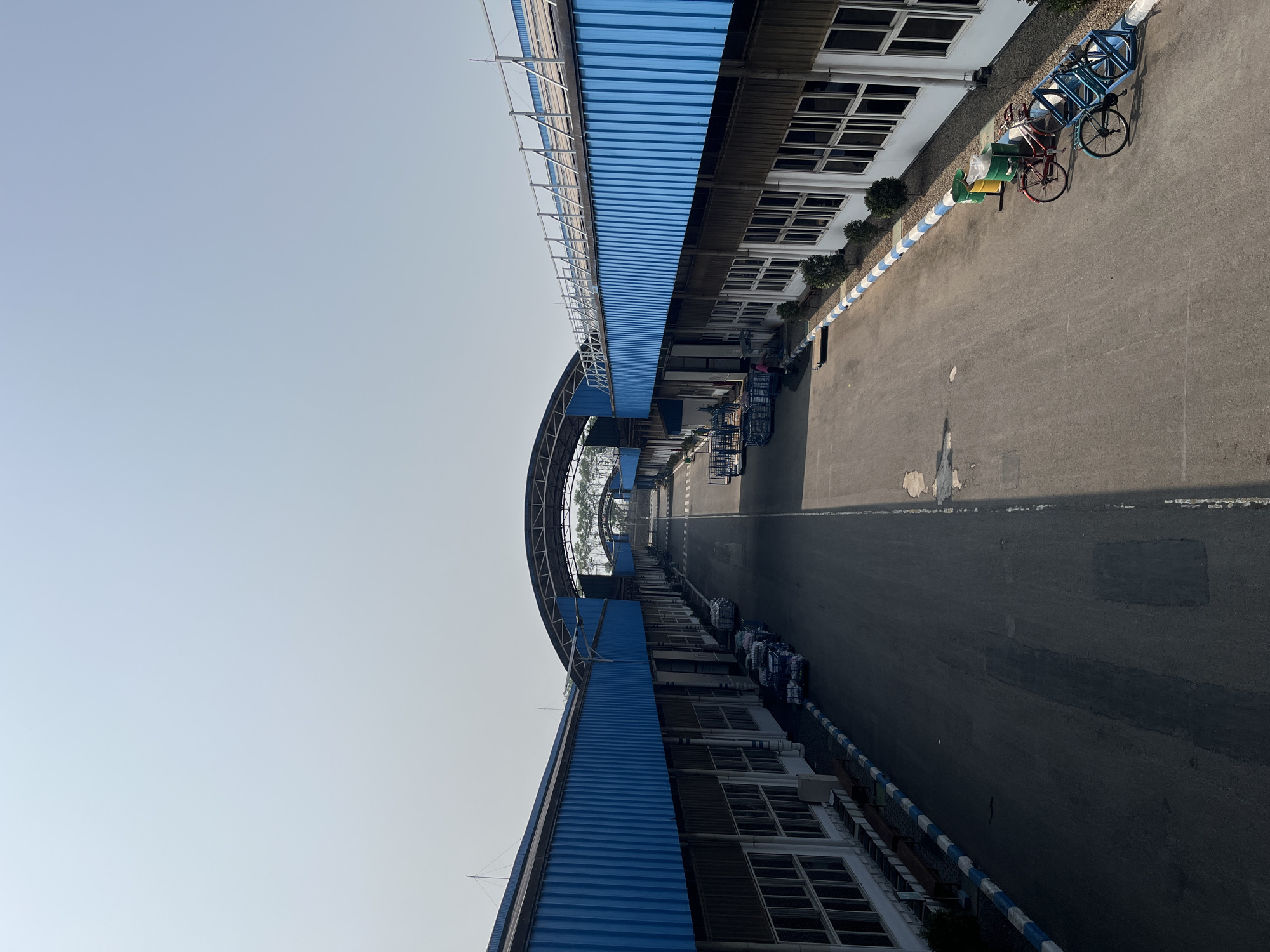 The image shows an aerial view of a large industrial complex with two long, parallel buildings on either side of a wide paved road. The buildings have blue corrugated metal roofs and white facades with numerous windows. A covered walkway with a curved roof structure connects the two buildings in the distance. Several carts loaded with materials are parked along the side of the road. In the foreground on the right side, there are two bicycles parked next to trash bins. The sky is clear and pale blue. The scene appears to be in a well-maintained industrial area, potentially a factory or warehouse complex. There are no discernible landmarks to pinpoint the specific location within Pelang, Indonesia.