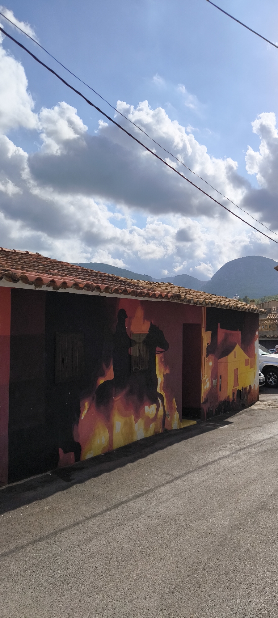 A street scene in Fanzara, Spain, features a mural on the side of a building. The mural depicts a silhouette of a person on horseback amidst flames and smoke. To the right of the horse and rider, the mural transitions to an artistic representation of buildings, some of which are illuminated with yellow and orange hues, suggesting either sunlight or fire. The building itself has a red-tiled roof and a dark-colored wall on the left side, which forms part of the mural's dark background. Wooden shutters are visible on the left side of the building. In the background, a white SUV is parked, and a hilly landscape with vegetation and some houses is visible under a sky with scattered clouds. Power lines stretch across the upper portion of the image. The perspective is from a street level, looking towards the building and the distant landscape.