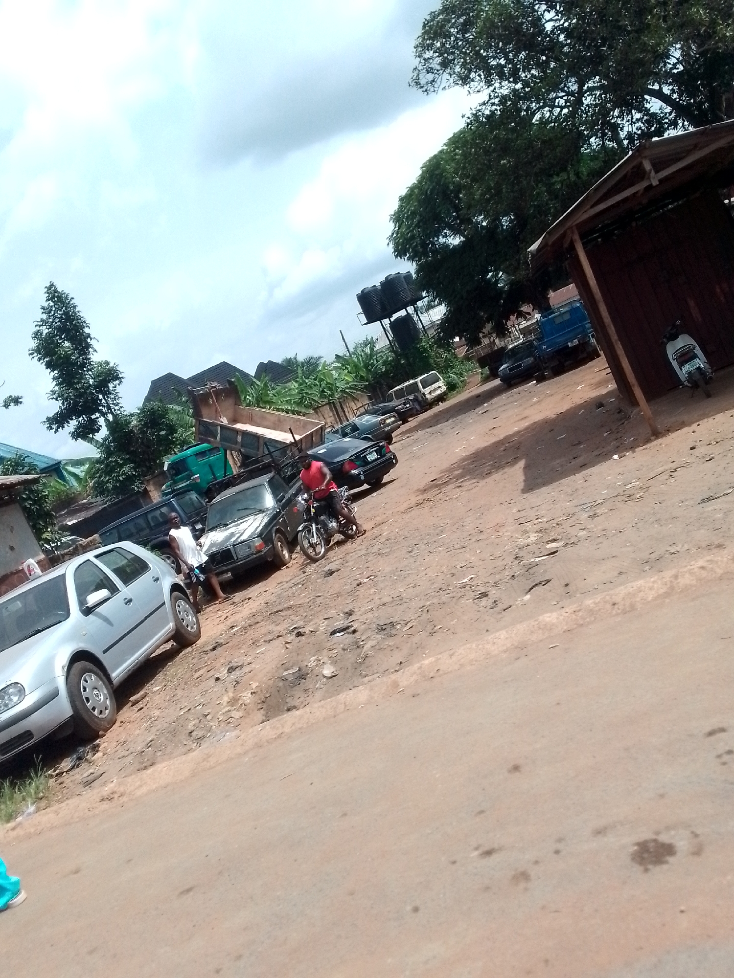 The image depicts a street scene in an urban area of the city of Unknown, Unknown. The street is mostly unpaved, dusty, and slightly inclined. There is a silver-colored car parked on the left side of the road. A small group of people, including one person on a motorcycle, are clustered near another parked car. Various other vehicles are scattered further back in the scene. A small wooden structure is located on the right side of the road, next to a parked scooter. Water tanks are visible in the background, as well as trees and buildings. The sky is partly cloudy.