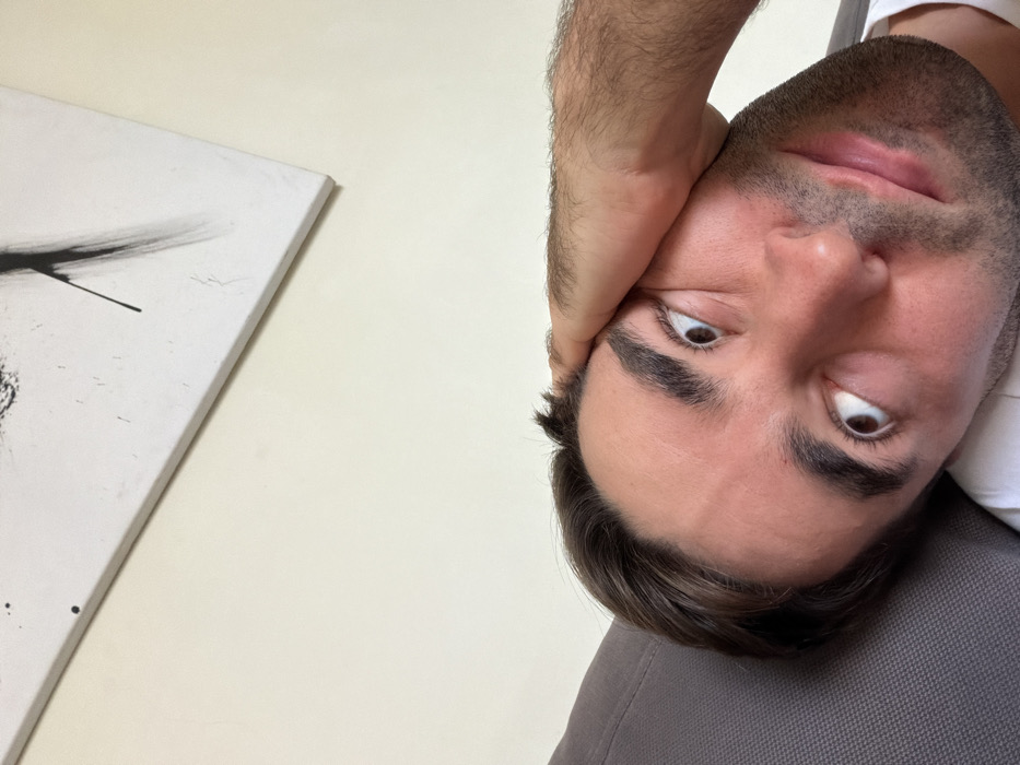 A close-up, low-angle indoor photograph displays an adult male reclining. His head, tilted to the left, rests against a textured gray upholstered surface. The male subject has fair skin, dark brown hair, prominent eyebrows, and visible stubble on his jawline and upper lip. His eyes are wide, directed upward and slightly to the right, accompanied by a subtle smile. His right hand, showing visible arm hair, is positioned against his forehead, partially obscuring his temple. A portion of a white t-shirt is visible at the bottom right. The background consists of a plain, light-colored wall, featuring a white rectangular canvas with abstract black brushstrokes and splatters mounted diagonally. The image contains no visual information to identify the city of Tel-Aviv, Israel.