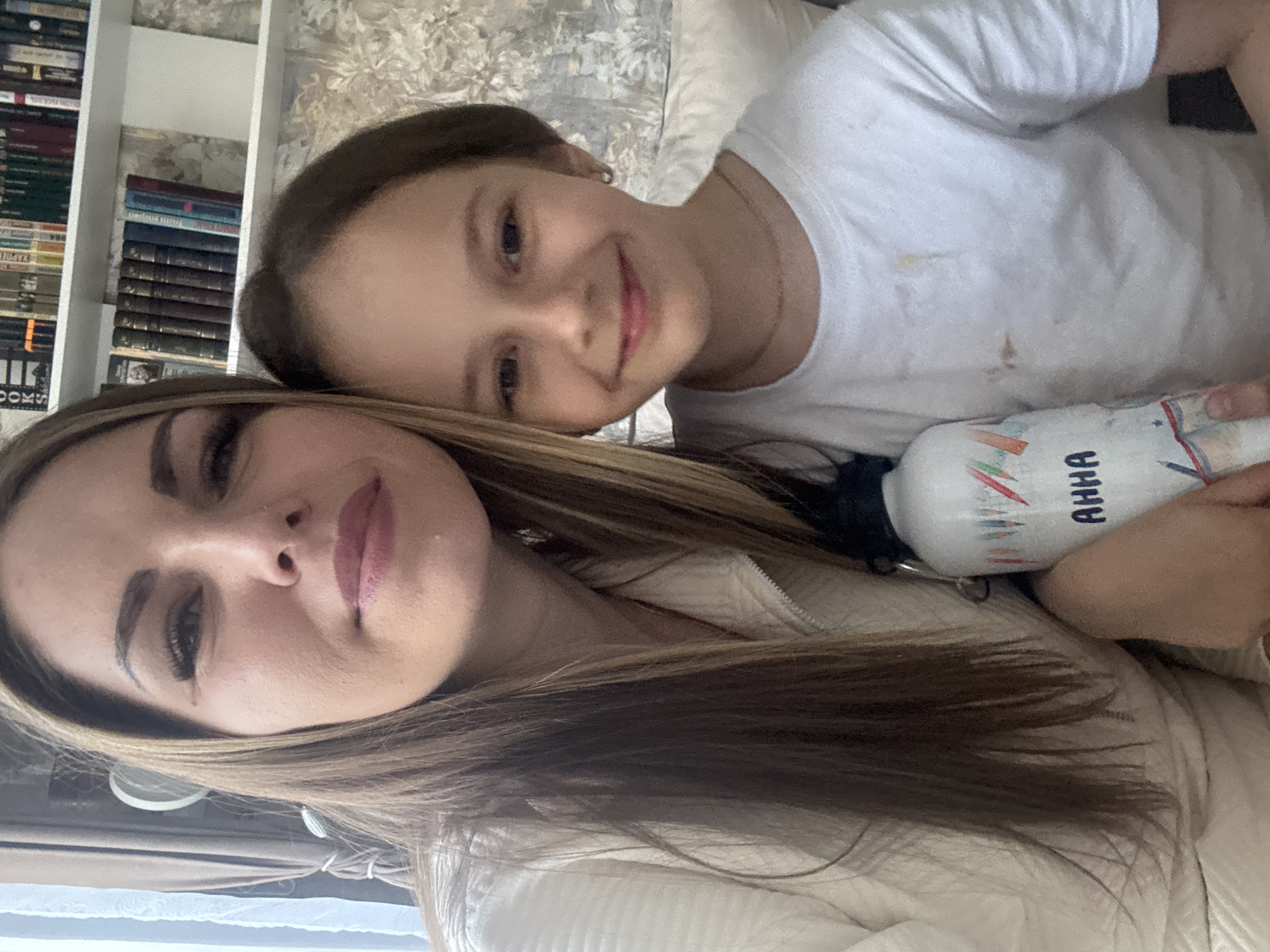 A selfie-style photograph shows two females positioned close together. The younger female, wearing a white t-shirt with what appears to be some stains on it, is situated behind the older female. She is smiling. The older female has long, light-brown hair and is wearing a cream-colored jacket. They are holding a water bottle together. In the background, there is a bookshelf and a wallpapered wall. The location is not discernible from the image.