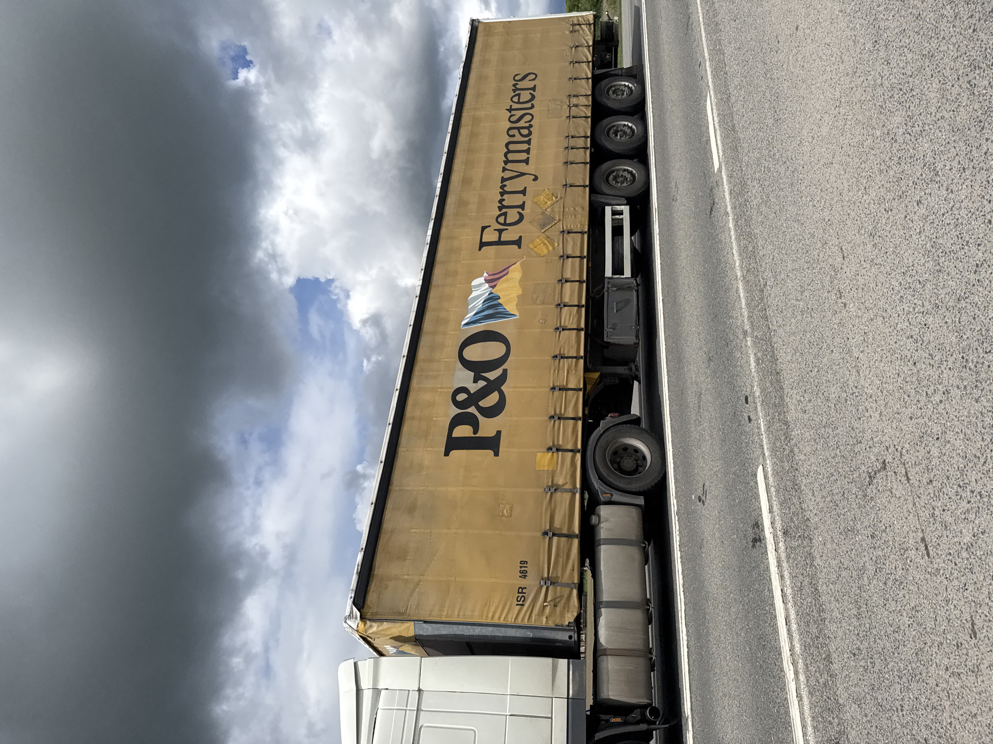 A white truck with a tan trailer that reads "P&O Ferrymasters" is traveling along a gray asphalt road. The sky above is partially obscured by a dark, ominous cloud formation. The location is not specified in the image and cannot be identified.