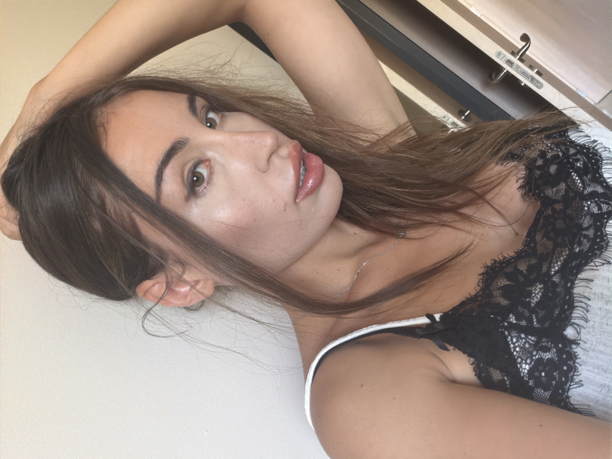 A medium shot shows a young woman with long, brown hair posing with her left arm raised behind her head. She is looking towards the camera with wide, brown eyes and glossy, pink lips. She is wearing a white tank top with black lace trim and a delicate silver necklace. The background is a plain, light-colored wall, and a portion of a wooden door with a metal handle is visible on the right side of the frame. The lighting is natural, suggesting an indoor setting, possibly a bedroom or dressing area, within a residence in Tersefanou, Cyprus.