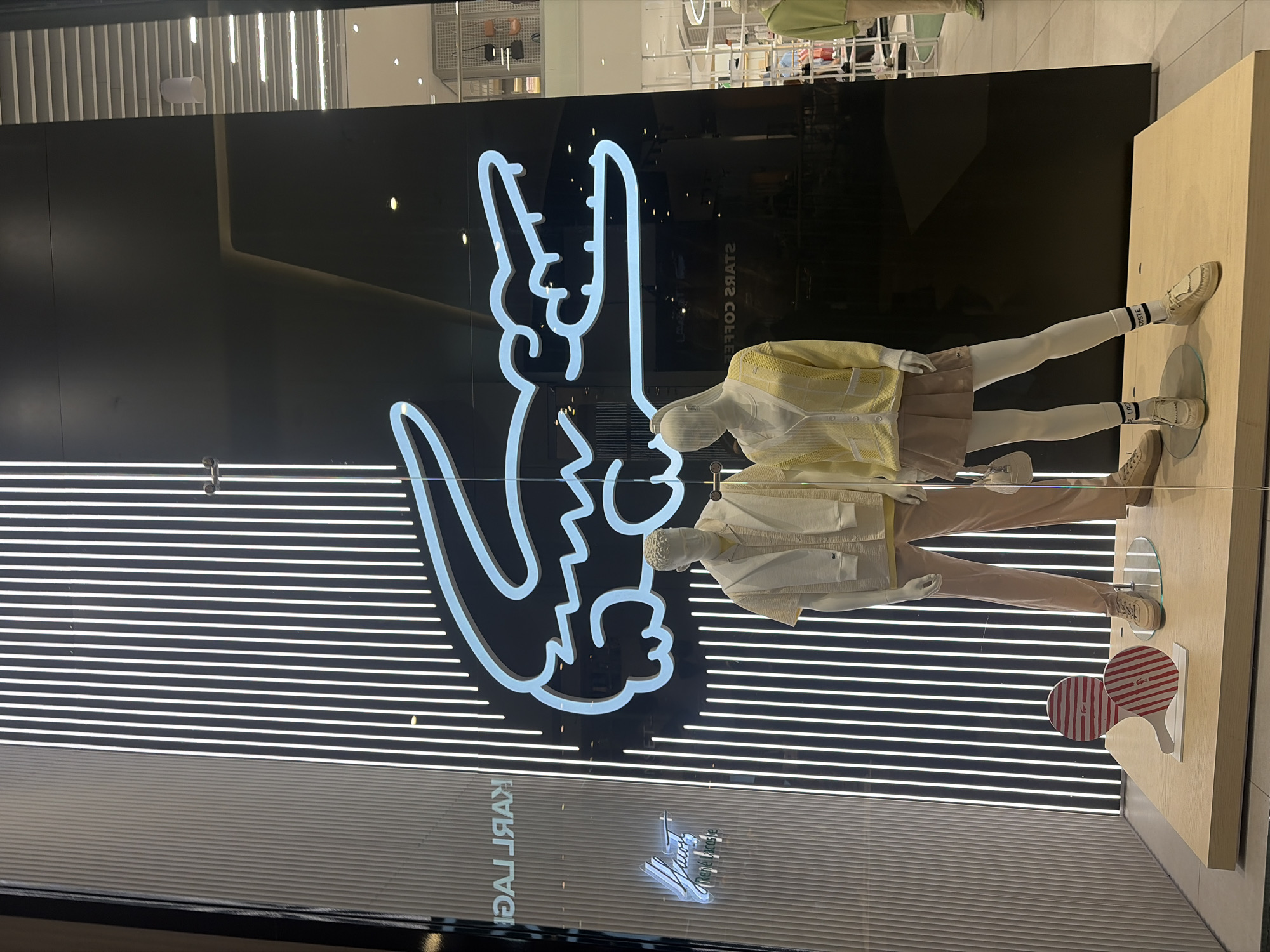 This image captures a retail shop window display, likely in Saint Petersburg, Russia, showcasing apparel and branding. The original image is rotated 90 degrees counter-clockwise.
The central focus is a large, illuminated, stylized neon sign on a black background, depicting the iconic Lacoste crocodile logo. Uniquely, the crocodile's head and tail are modified to form a peace sign or "V for victory" hand gesture, glowing in white.
In front of this background are two mannequins dressed in contemporary casual wear. The mannequin on the right wears a light yellow, textured cardigan over a white top, a light pink or beige pleated skirt, white tights, and light-colored loafers with black and white striped socks featuring the "LACOSTE" brand name. The mannequin on the left is dressed in a white textured jacket, a white top, light brown pants, and similar light-colored shoes. Both mannequins are positioned on a light wooden display platform.
Also on the wooden platform, next to the mannequin on the left, are two decorative, paddle-shaped objects with red and white stripes, one bearing a small Lacoste crocodile logo.
The shop window reflects elements from the interior and exterior environment. Visible reflections include the reversed text "STARS COFFEE" and "KARL LAGERFELD," suggesting other stores or signs nearby. Another small, reflected Lacoste logo is also visible below the large peace-sign crocodile. The lighting suggests an indoor setting, likely a shopping mall, with horizontal light strips visible in the background reflection.