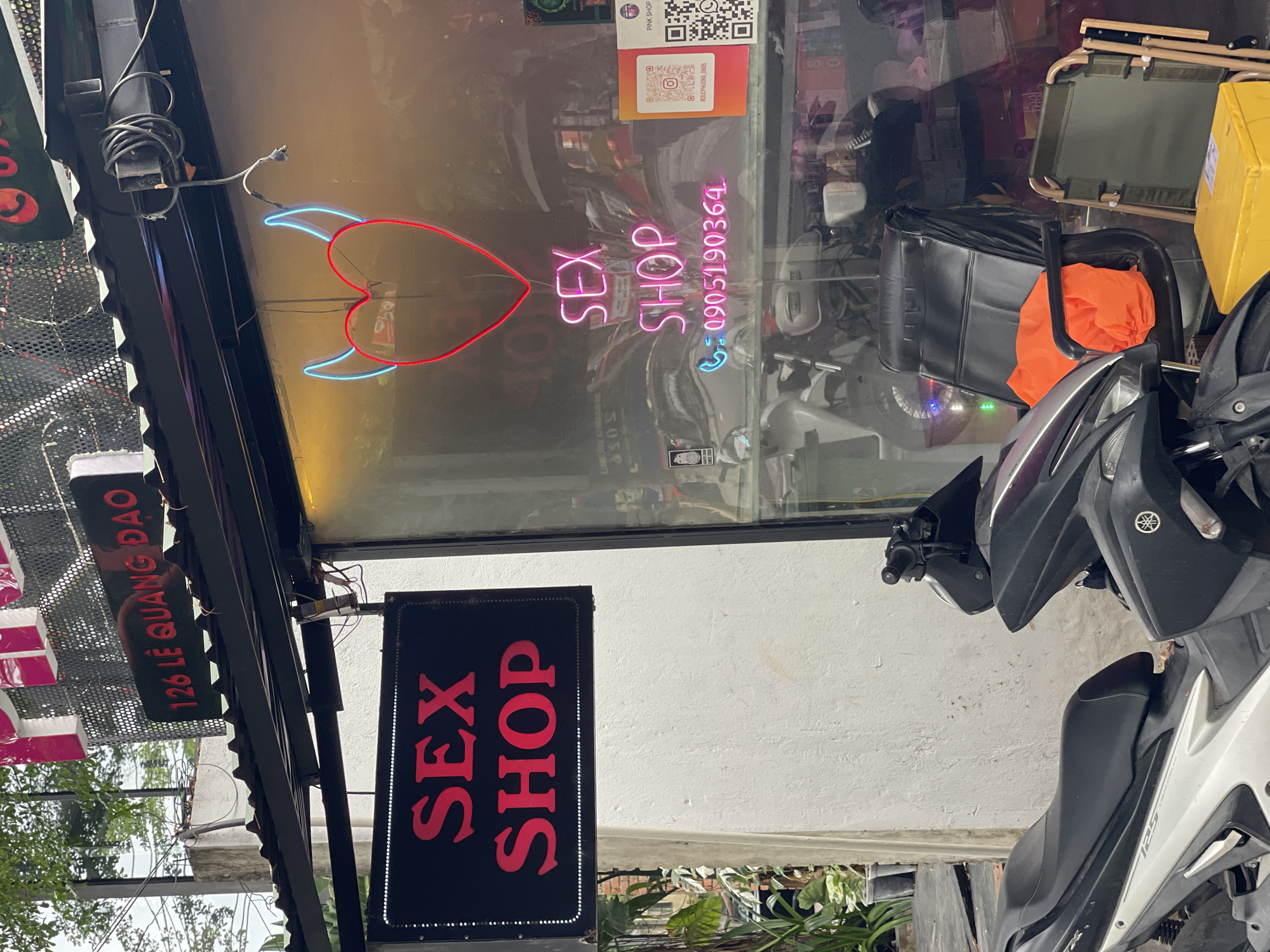 The image depicts the storefront of a sex shop located at 126 Lê Quang Đạo street in the city of Đà Nẵng, Vietnam. A prominent red neon sign spelling "SEX SHOP" in English is displayed on a black background, hanging to the left of the main entrance. Inside the shop window, a neon sign forming a red heart with blue devil horns is visible, along with another "SEX SHOP" sign in pink neon. Below the pink neon sign, a phone number "0905190364" is illuminated in blue neon. Two motorcycles are parked in front of the shop, and a black office chair with an orange bag draped over it, along with a folded green camping chair, are placed near the entrance. The building exterior is a white wall, and a corrugated metal roof extends over the shopfront. Trees and greenery are visible in the background.