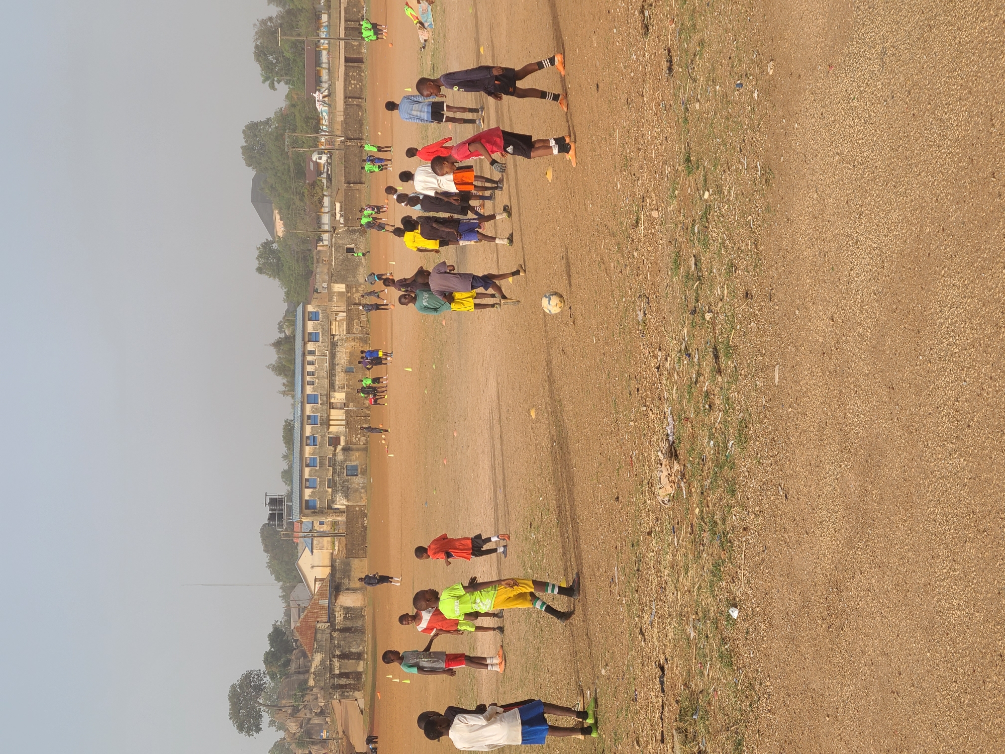 The image depicts a soccer field in Jos, Nigeria, where a group of people are engaged in athletic activity. The field surface appears to be predominantly bare earth and dry grass, and is surrounded by a perimeter of grassy vegetation. Scattered debris is seen along the edges of the field.
Numerous individuals are spread across the field, some appearing to be performing push-ups or stretching exercises, while others are in other training positions. The individuals' attire consists of various sportswear items, in different colors, including green, yellow, red, blue, and black. A soccer ball rests on the field among the people.
A cluster of low-rise buildings is visible in the background, including a prominent multi-story building with blue-framed windows. Trees and foliage surround the buildings and stretch across the skyline, with a hazy sky overhead.