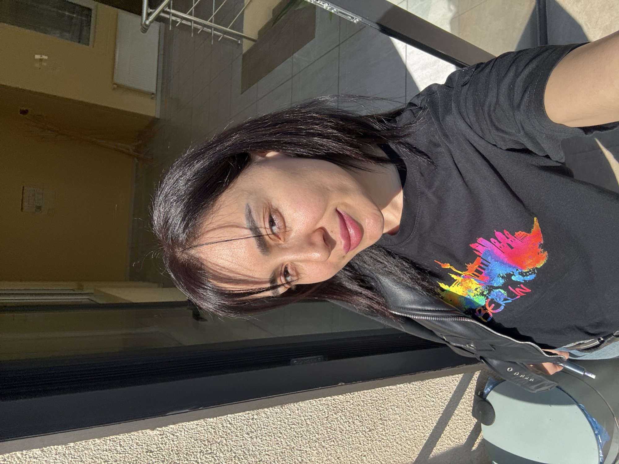 A woman stands on a balcony of a building, likely in Tbilisi, Georgia, based on the "Berlin" text and city skyline depicted on her black t-shirt. She wears a black leather jacket and appears to be holding a selfie stick attached to a round, light blue object, perhaps a phone holder or stand. The balcony overlooks an exterior space with light-colored tile flooring and an interior space visible through a window. The light indicates it's daytime.