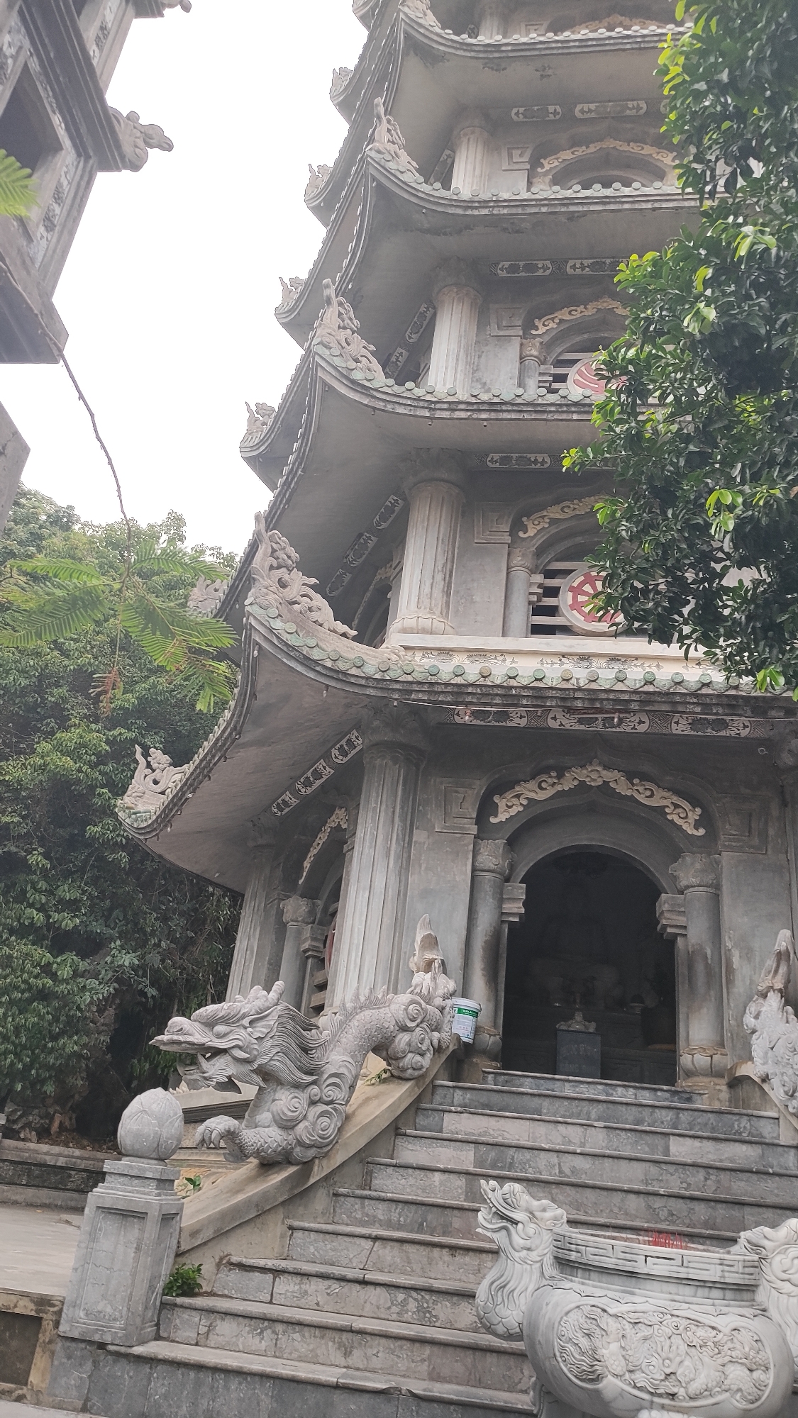 The image shows a multi-tiered pagoda, characteristic of Vietnamese architecture, with grey concrete construction. Intricate dragon carvings adorn the railings of the ascending staircase. The pagoda features curved eaves with decorative elements and pillars. Trees and foliage surround the structure, suggesting an outdoor, possibly temple or garden setting, located in Đà Nẵng, Vietnam. The sky is overcast, casting a diffused light on the scene.