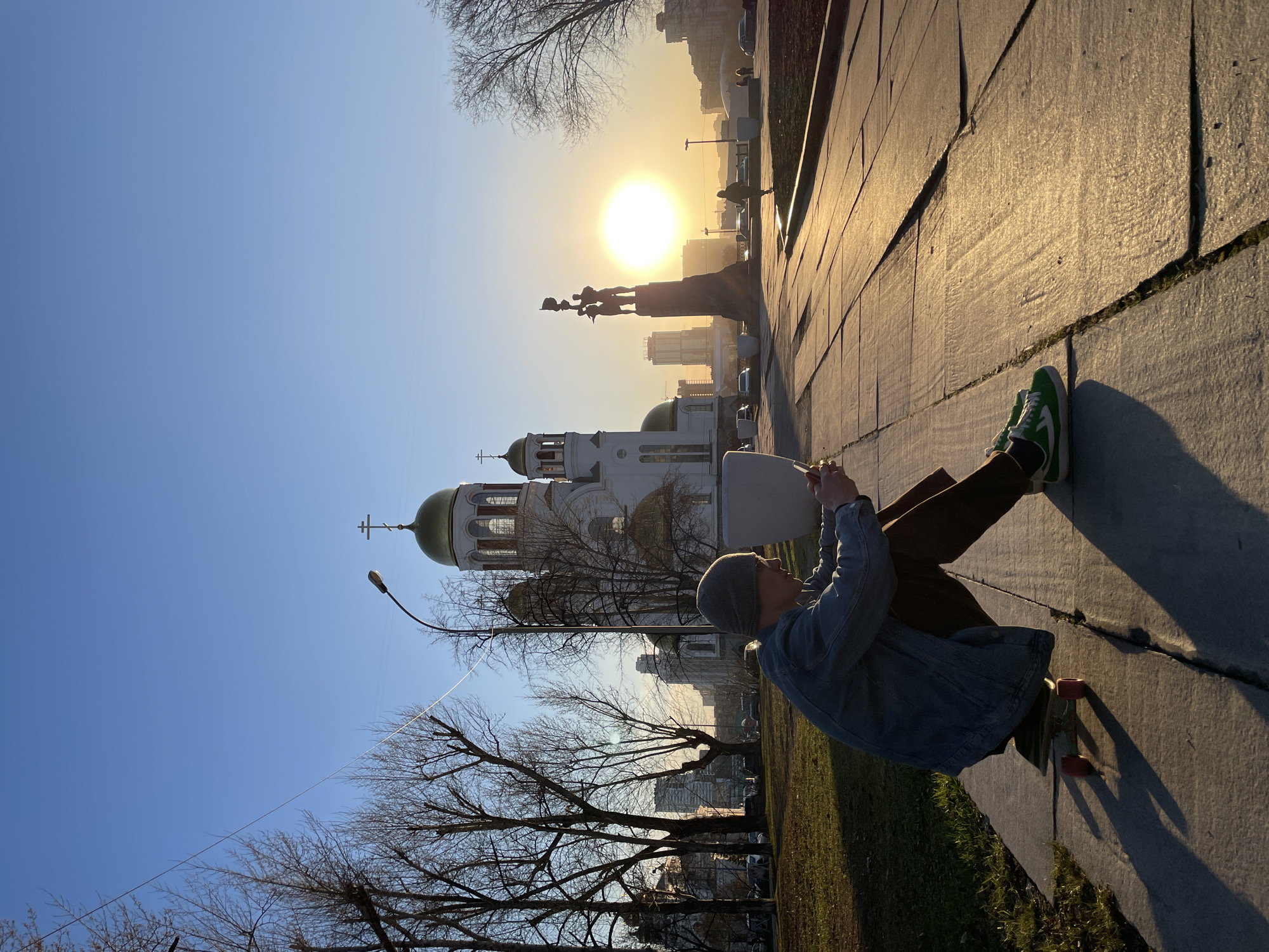 A person sits on the edge of a sidewalk, using a phone to take a photo of the Church on Blood in Yekaterinburg, Russia. The monument to the founders of the city is visible in the background, with the sun behind it. The person is wearing a denim jacket, brown pants, green and white sneakers, and a gray beanie. A skateboard lies behind them. Bare trees stand between the person and the church, and a street lamp is positioned to the left of the church. The sky is clear and blue.