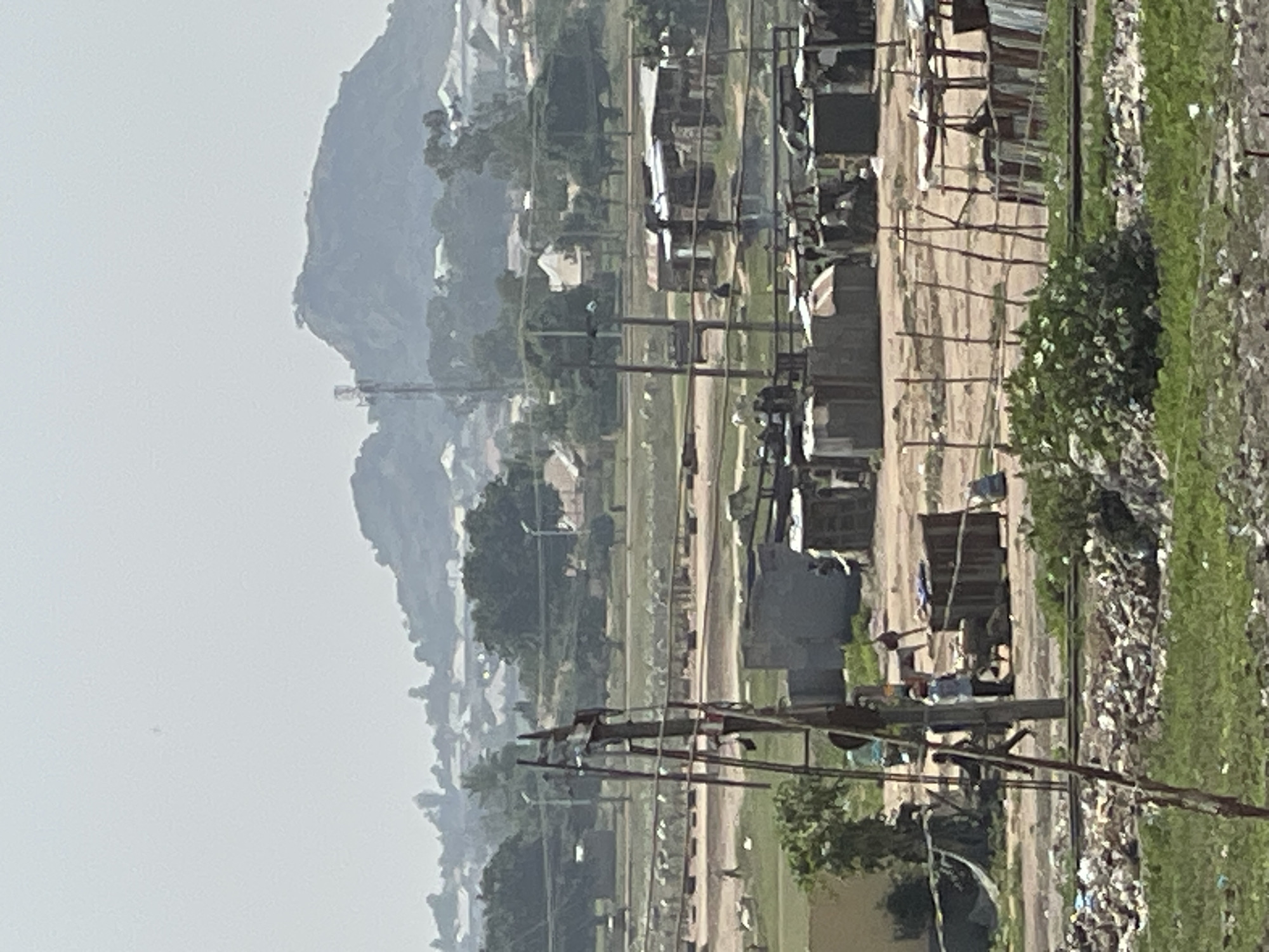 The image depicts a view in Bukuru, Nigeria, showing a landscape that includes a hill or mountain in the background, shrouded in haze. In the foreground, there are structures resembling shacks or informal settlements lining a dirt road. Power lines and utility poles run along the road. There is vegetation present, with what appears to be overgrown grass and some bushes. Debris and possibly trash can be seen interspersed with the vegetation. The image suggests a location on the outskirts of a developed area, bordering a less formal settlement. There are no people visible in the image.