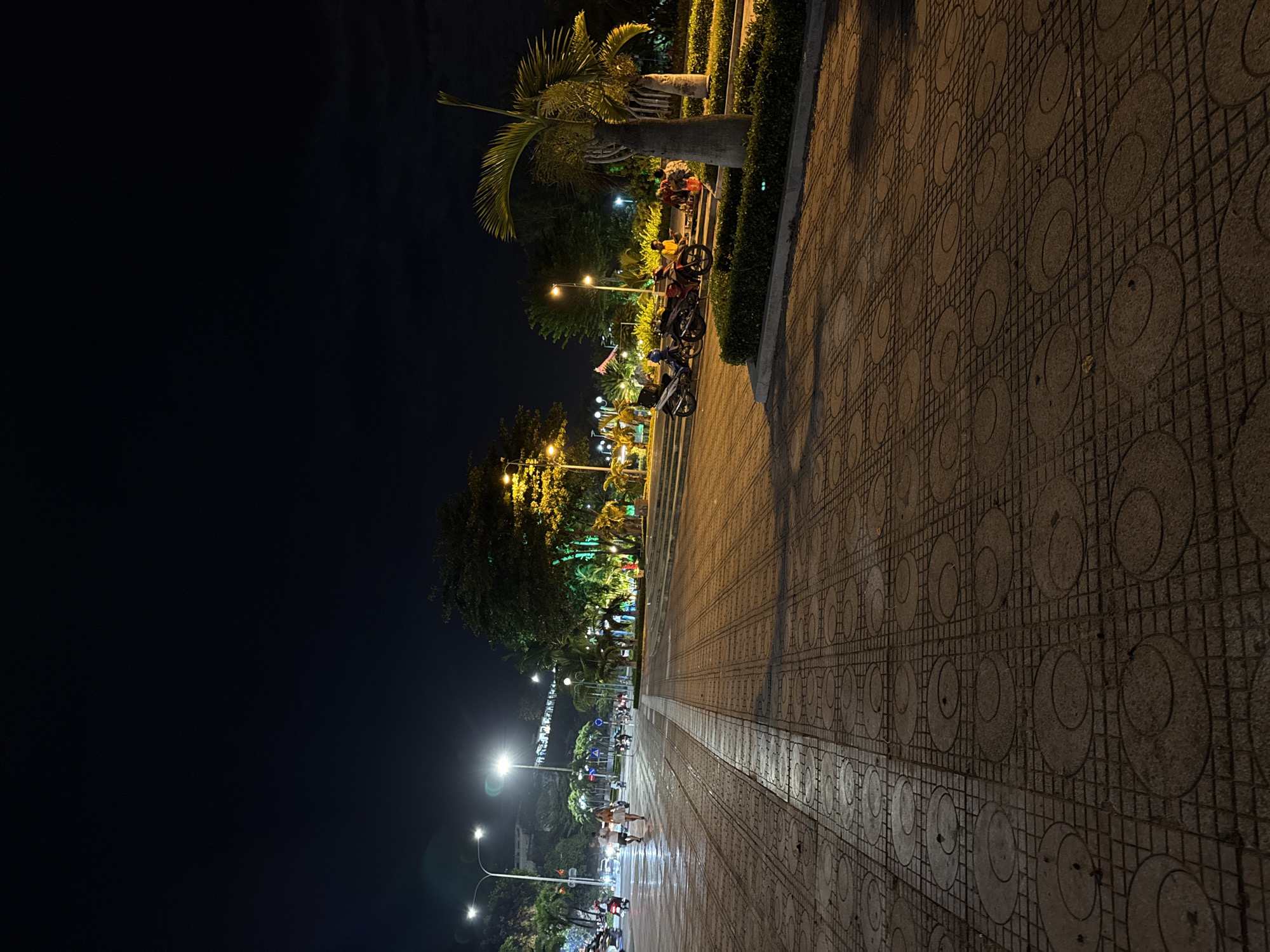 The image depicts a wide, paved walkway at night in Nha Trang, Vietnam. The walkway is made of square tiles, with a section featuring circular patterns. Streetlights illuminate the scene, casting long shadows. On the left side of the walkway, a street is visible with several motorcycles driving by. In the background, there are trees and greenery, some of which are lit with colorful lights, suggesting a park or public space. A group of people are gathered near some parked motorcycles on the right side of the walkway, seated and interacting. The overall atmosphere is urban and lively, typical of a night scene in a Vietnamese coastal city.