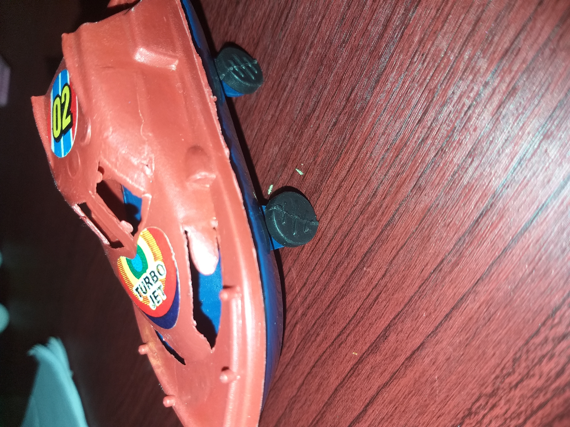 A red plastic toy airplane with a blue bottom and black wheels is resting on a dark reddish-brown wooden surface. The toy has stickers on it; one depicts a rainbow with the words "TURBO JET" and another has the number "02" within a circle. The toy appears to be on a flat surface, possibly a table or floor. The background is out of focus, but suggests an indoor setting. There are no people visible in the image. The overall scene is a close-up shot of the toy airplane. There is no information in the image to suggest a specific location within Air Jamban, Indonesia.