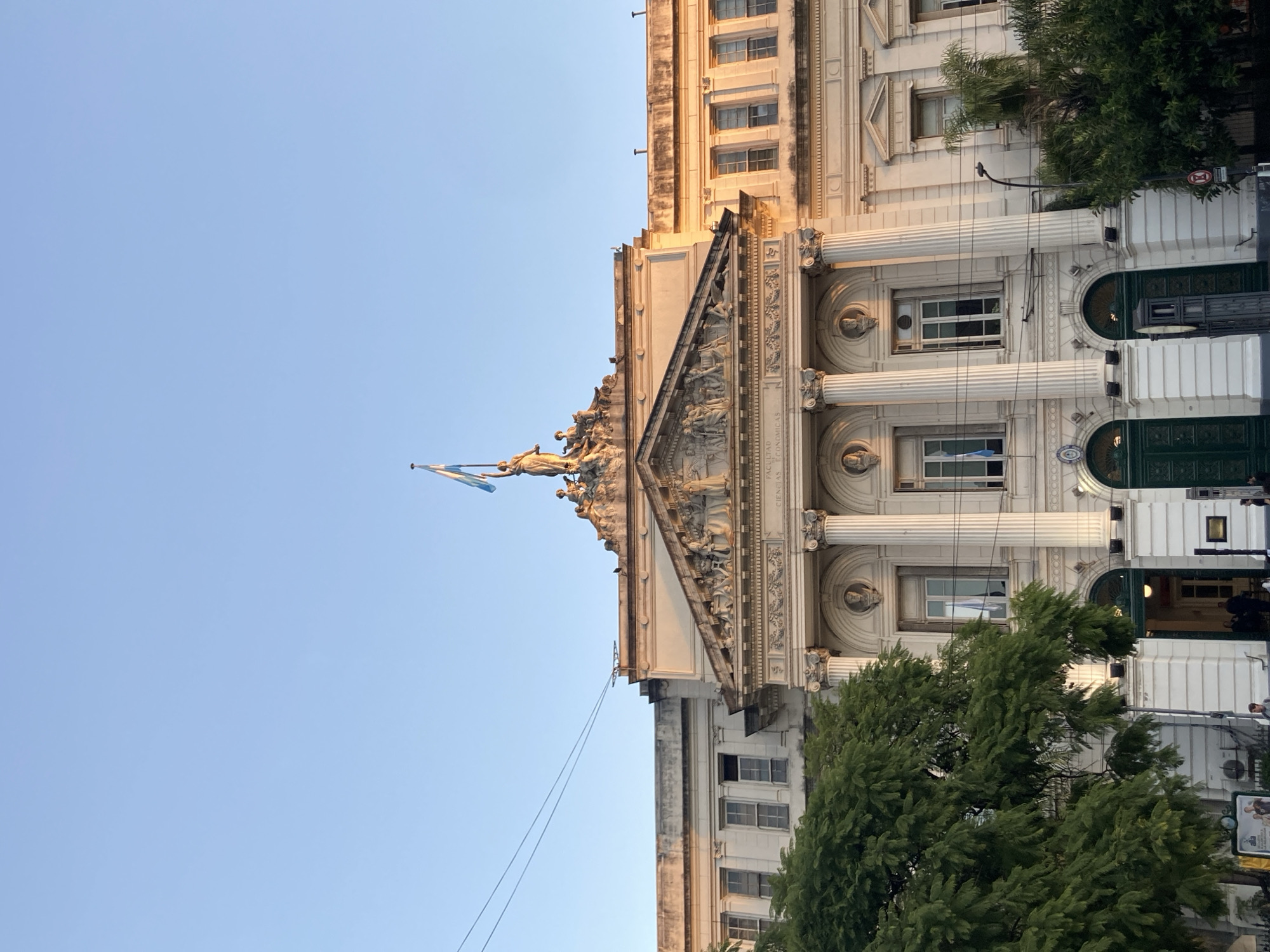 The image captures a grand, classical-style building, identified as the "FACULTAD DE CIENCIAS ECONÓMICAS" (Faculty of Economic Sciences), under a clear, light blue sky. The architecture features multiple stories, prominent fluted columns with ornate capitals, and intricate decorative carvings.
Atop the building's central pediment, a prominent sculpture group includes a standing figure holding a flag, which is clearly identifiable as the flag of Argentina with its light blue and white stripes. The upper facade of the building and the statue are bathed in warm sunlight, suggesting either late afternoon or early morning.
The lower portion of the building is partially obscured by dense green trees. Several windows are visible, and at ground level, there are large, dark green double doors. Scattered street furniture, including a red circular traffic sign and a public information board, can be seen through the foliage. Thin utility wires are visible against the sky. The scene is an urban landscape, consistent with the provided context of Buenos Aires, Argentina. No specific activity or individuals are prominently featured.