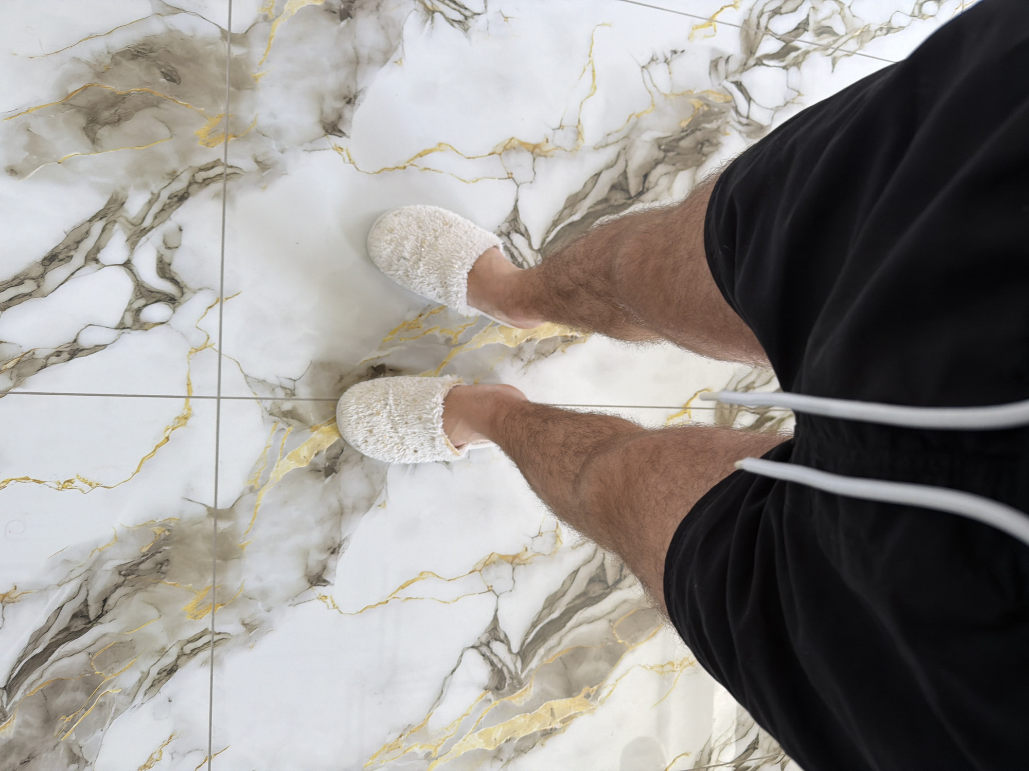A top-down perspective shot shows a person's legs and feet, clad in fluffy white slippers and black shorts, standing on a marble-patterned floor. The floor features prominent white and grey veining with thin, gold-colored accents, divided by thin grout lines. The individual's legs are covered in hair, and the black shorts have a white drawstring. The setting is indoors, likely a bathroom or living area, with no visible indication of the specific location within Strășeni, Moldova.