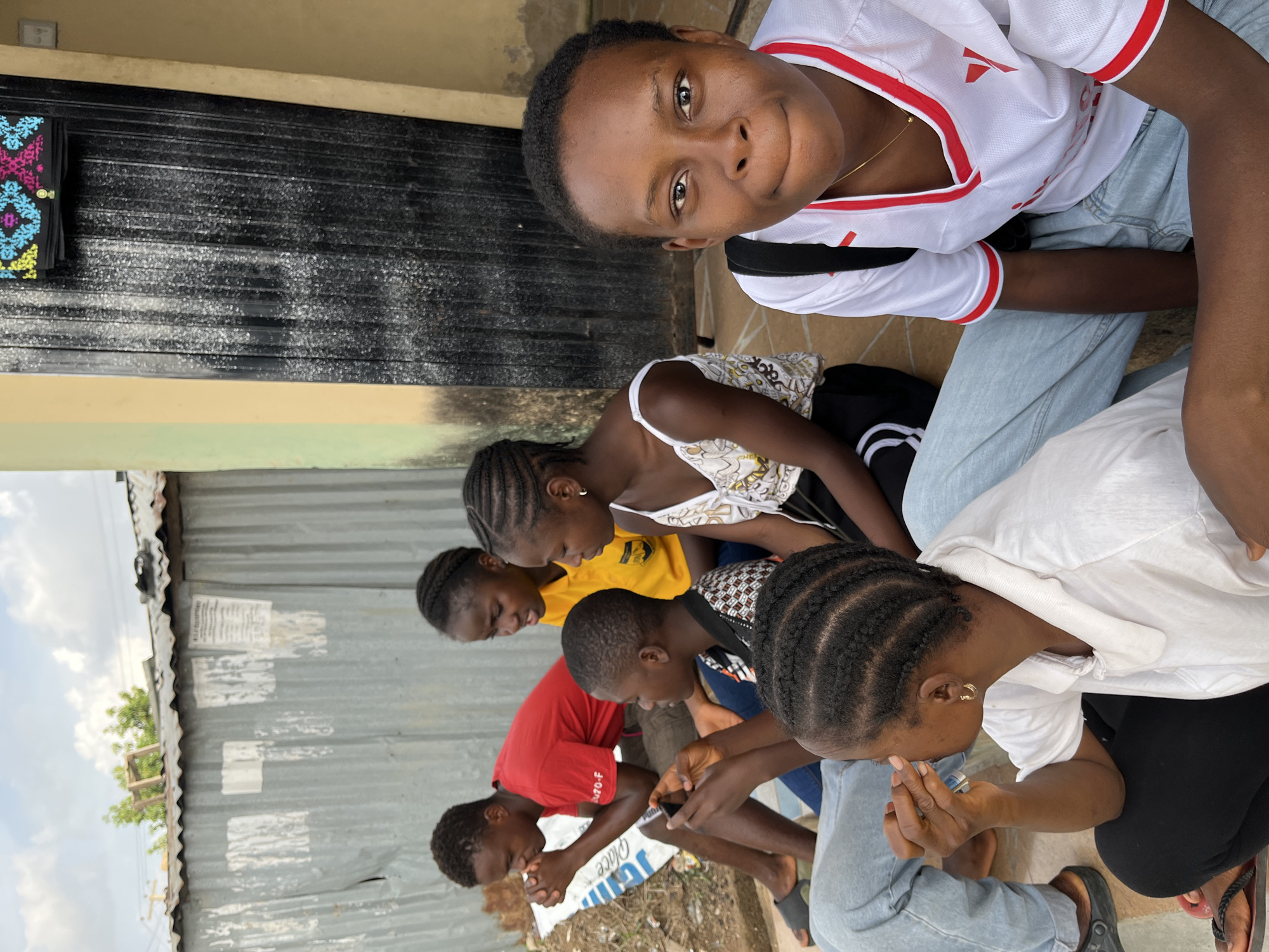 A group of seven individuals, predominantly young, are seated on the ground outside a building in what appears to be Jos, Nigeria. One person, wearing a white shirt with red accents and light blue jeans, is looking upwards at the camera. Another is wearing a white tank top with a pattern and black pants. The rest of the group are wearing yellow, red, and white shirts. Most of the group are focused on items in their hands. The background includes a corrugated metal fence and a black, metal door. Trash and debris are scattered on the ground.