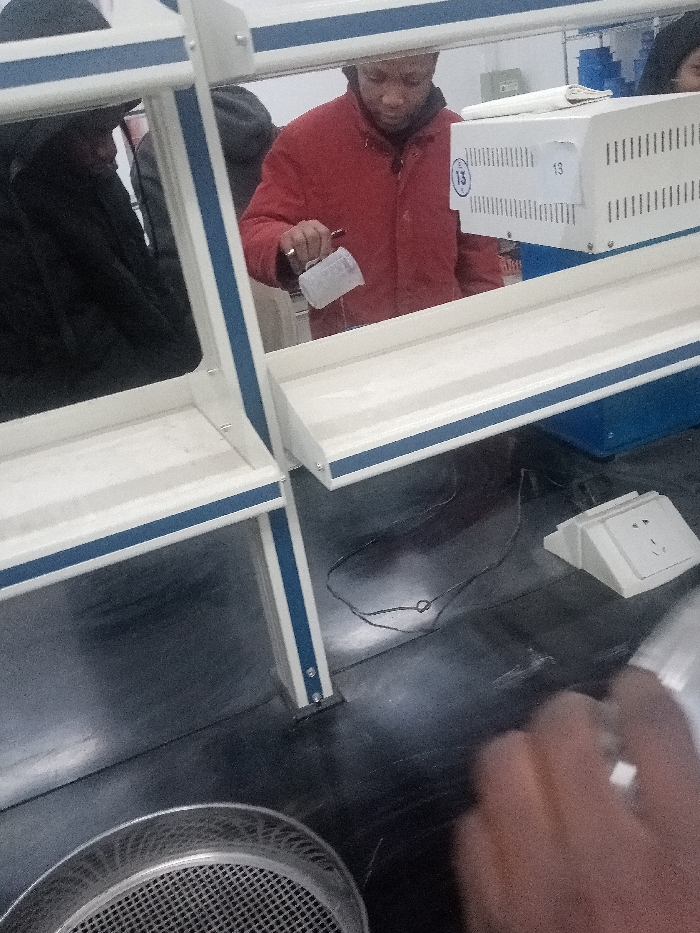 A person in a red jacket holds a plastic container over a metal shelf, possibly pouring liquid. The shelf is part of a white and blue structure with an electrical outlet nearby. A sieve sits on the dark surface below. The setting appears to be a laboratory or workshop environment. Another individual is partially visible on the left.