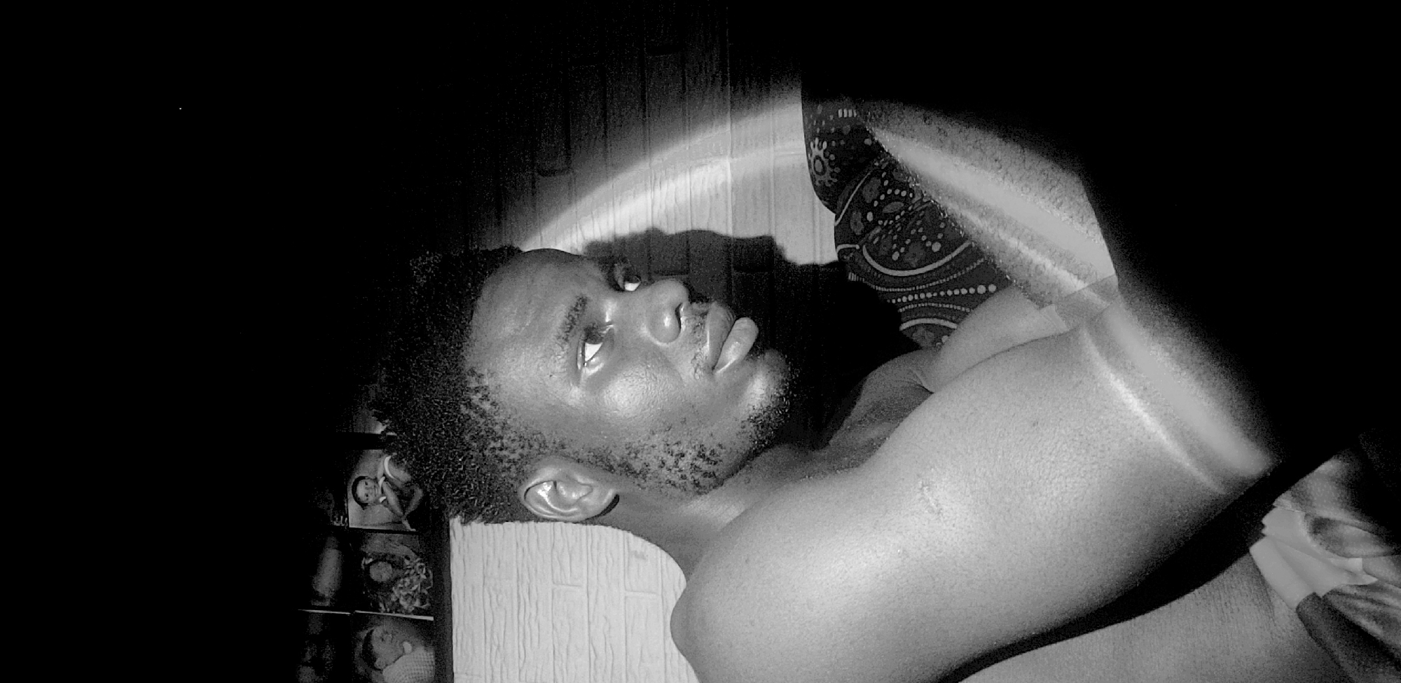 This black and white portrait features a young man lying down in what appears to be an indoor setting in Narayi, Nigeria. The man, with dark skin, short curly hair, and light stubble on his face, is shirtless or wearing minimal clothing on his upper body. He is positioned on his side, looking upwards and slightly away from the camera, with a contemplative or relaxed expression.
The primary light source is artificial, creating a striking, bright, curved arc across the textured wall behind his head and shoulder. This dramatic lighting casts his face and upper body in strong highlights and shadows, emphasizing his features and the texture of his skin, which appears somewhat dewy. The rest of the room is largely plunged into darkness, creating a moody and intimate atmosphere.
To the right of the frame, a patterned fabric, possibly a pillow or blanket, is visible. On the far left, in the dimly lit background, there's a shelf or display unit holding several small framed photographs, one of which clearly depicts a child. The man's posture and gaze suggest a moment of quiet reflection or repose, captured in a personal and artistic manner. No visible text is present in the image.