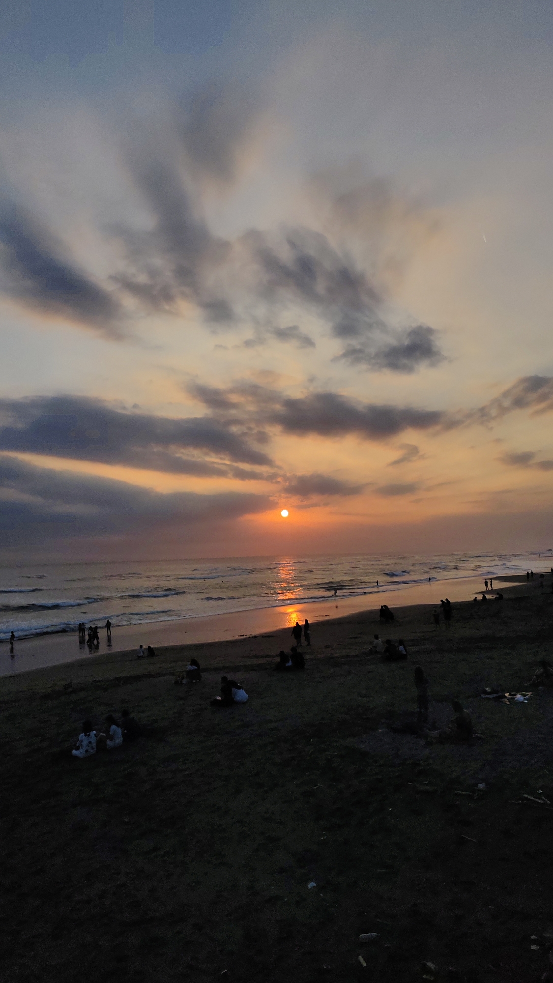 The image captures a serene sunset scene at a beach, likely in the city of Kediri, Indonesia, given the context provided. The sky is a gradient of soft blues and oranges, with streaks of gray clouds adding texture and depth. The sun, a vibrant orange orb, is setting near the horizon, casting a shimmering golden reflection on the calm sea. Gentle waves lap at the sandy shore, where a scattering of people are visible. Some individuals are standing near the water's edge, while others are seated or lounging on the dark sand, enjoying the twilight. The foreground is dominated by the dark, textured sand of the beach, with a few scattered objects that are indistinguishable due to the low light. The overall atmosphere is peaceful and tranquil, typical of a beach setting at dusk.