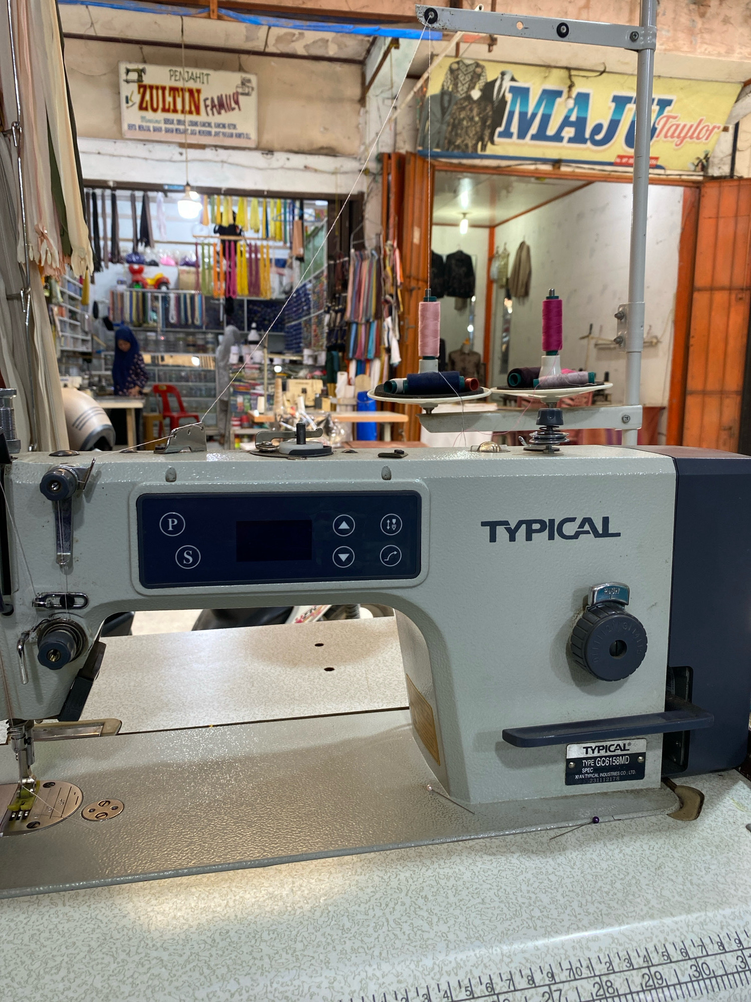 The image shows a close-up view of a sewing machine in what appears to be a tailor shop in Lhokseumawe, Indonesia. In the background, signs indicate the names of tailoring businesses, "ZULTIN FAMILY" and "MAJU Taylor". The shop is stocked with various fabrics and threads, suggesting an active tailoring business. The sewing machine itself is modern, with a control panel featuring buttons and a digital display. Several spools of thread are visible on the machine and in the background. The overall impression is of a working tailor shop environment.