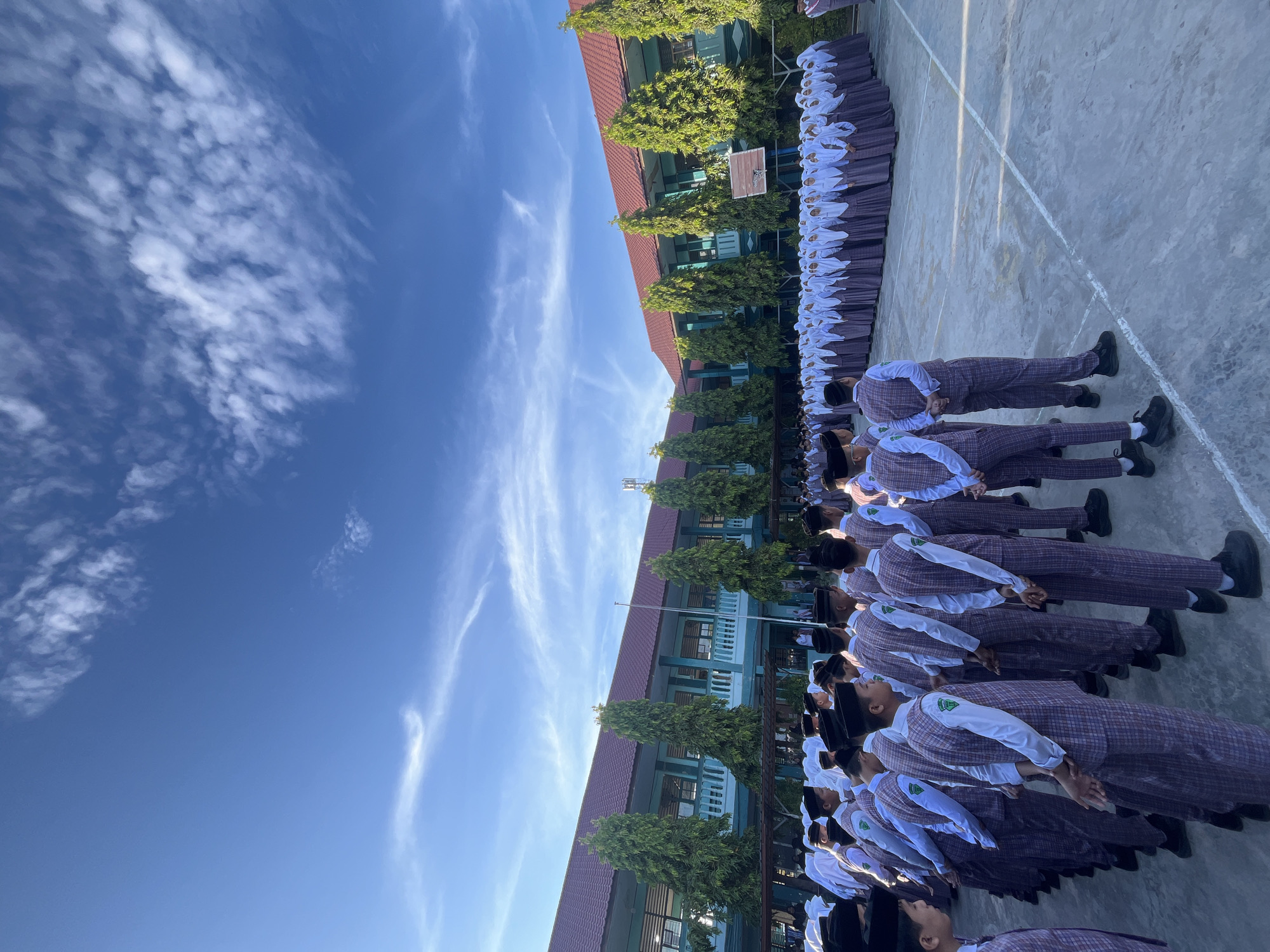 A wide-angle, eye-level shot shows a group of male students in a schoolyard, facing away from the camera and towards a building. The students are wearing plaid uniforms with white shirts and dark caps. They are lined up in formation on a paved surface with white lines marking areas. Behind the students is a two-story building with a red roof and light blue walls, flanked by tall, slender green trees. The sky above is a clear blue with wispy white clouds. The scene suggests a school assembly or event taking place outdoors in Lhokseumawe, Indonesia, likely during daytime.
