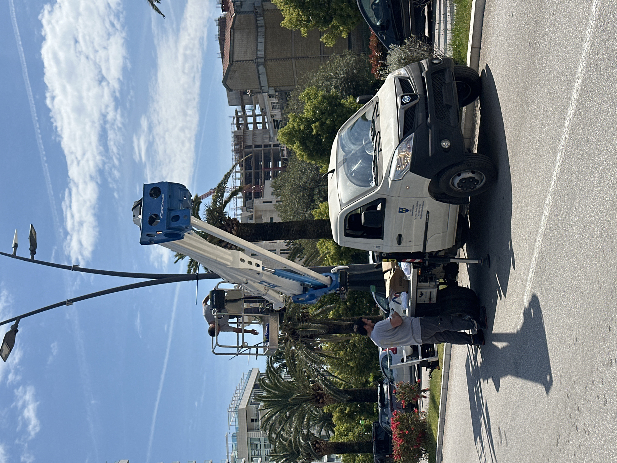 This image captures an outdoor scene of utility work in Budva, Montenegro, under a clear, sunny sky with scattered wispy clouds. A white service van, possibly a Renault Kangoo, is parked on the asphalt road, serving as the base for a white and blue articulated boom lift (cherry picker).
Two workers are engaged in the activity. One worker, wearing a grey t-shirt and dark pants, is elevated in the boom lift's basket, positioned near a street light. Another worker, also in a grey t-shirt and dark pants, stands on the ground by the rear of the van, looking up towards the basket, likely overseeing or operating the equipment. Strong shadows cast on the road indicate bright midday or early afternoon sunlight.
In the background, several buildings are visible, including a large structure under construction with exposed concrete frames. Finished buildings, possibly apartments or hotels, are also present. The scene is characterized by abundant green foliage and several tall palm trees, suggesting a Mediterranean or coastal environment.
Notable visible text includes "COCAGE" on the blue part of the cherry picker's basket. The side of the white van features a blue shield-like logo with white text, and further unreadable text, likely an organization's name or contact information. The front of the van displays a diamond-shaped logo on its grille.