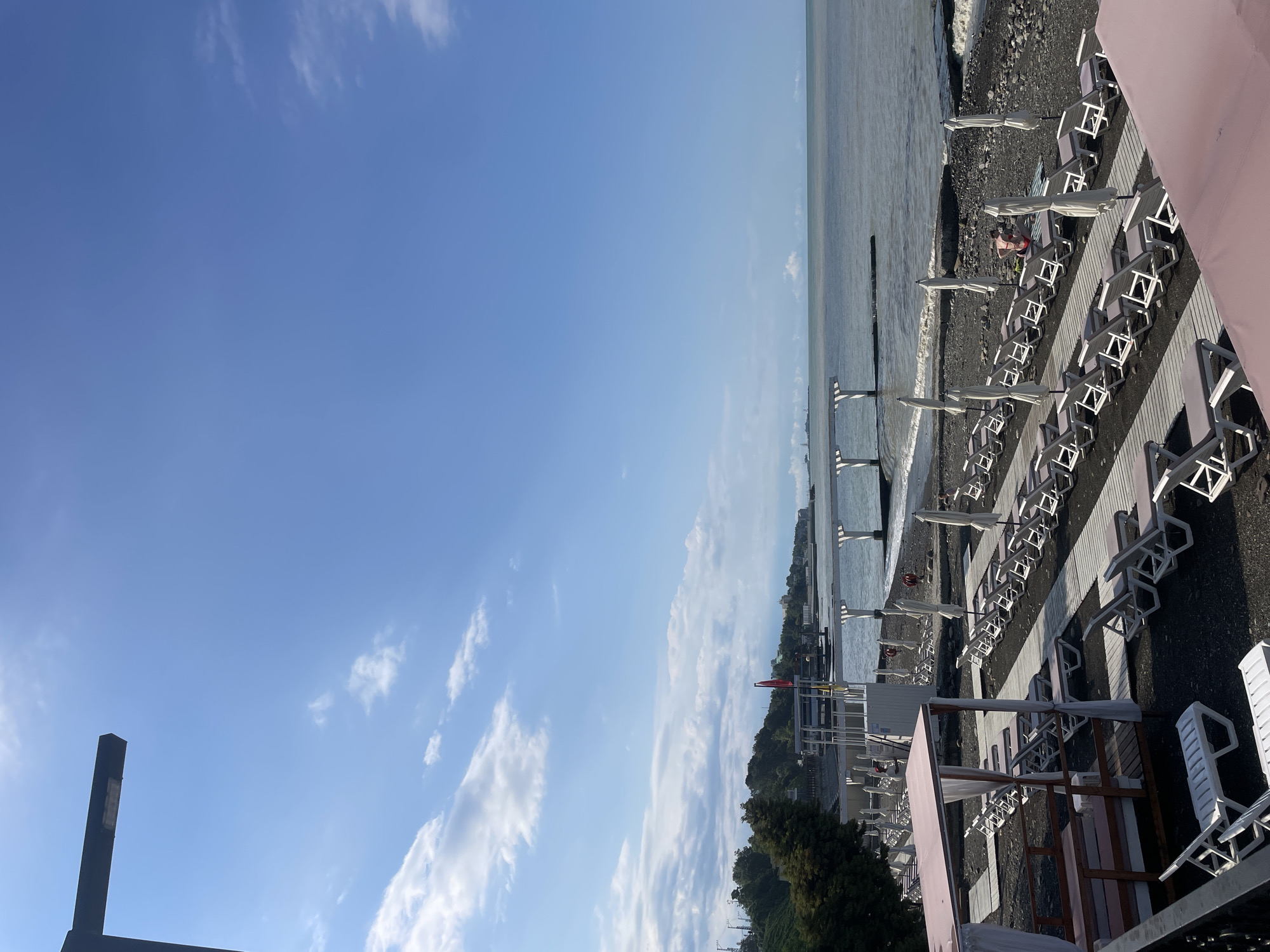A wide shot of a beach in Sochi, Russia. The beach is covered in dark pebbles and lined with rows of empty white lounge chairs and closed white umbrellas. Wooden walkways lead across the beach to the water. The sea is a muted gray with small waves. In the distance, there is a breakwater and several white structures over the water. The sky is blue with scattered white clouds. A few trees are visible on a hill behind the beach. A single person wearing a red swimsuit is sitting on a lounge chair near the water.