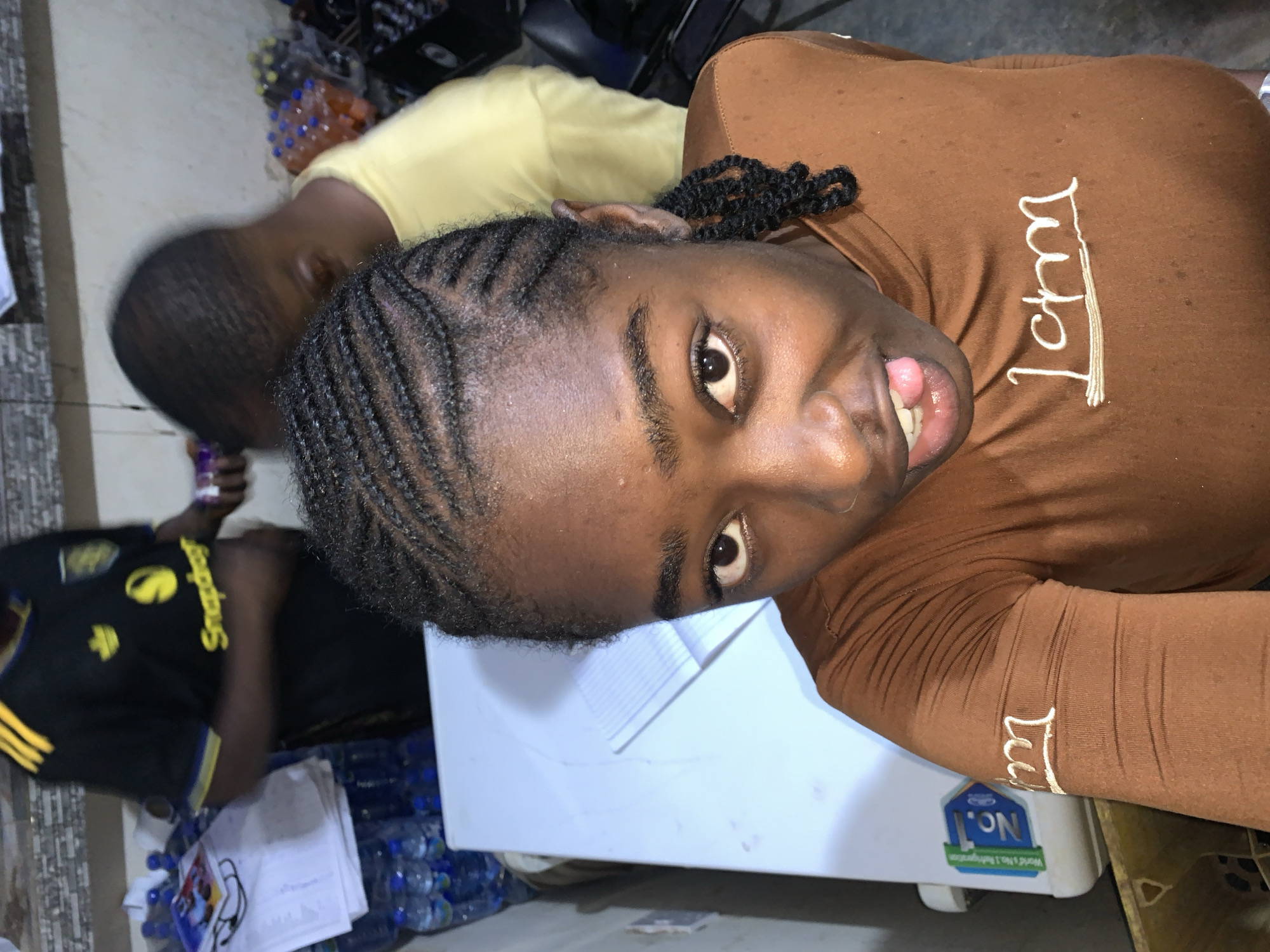 The image captures an indoor scene, likely a commercial establishment or a storage area. In the foreground, a dark-skinned individual with dark braided hair (cornrows) faces the camera directly, holding their tongue slightly extended. They are wearing a brown long-sleeved top with white embroidered lettering across the chest that appears to read "I Am," and similar embroidery on the right sleeve.
Behind the foreground subject, two other individuals are partially visible and out of focus. To the left, a person wearing a black short-sleeved shirt with yellow stripes and a "Snapdragon" logo is standing behind a white table or counter. On this table, a stack of papers and numerous plastic bottles of water are present. To the right, another individual wearing a light yellow short-sleeved shirt is visible, largely obscured by the foreground subject, and appears to be looking downwards. In the far right background, additional stacks of bottled items are visible. The background wall features a textured, possibly tiled, surface. The lighting suggests an indoor setting with artificial illumination. The precise location, including the city of New Stadium, Nigeria, is not discernible from the visual evidence.