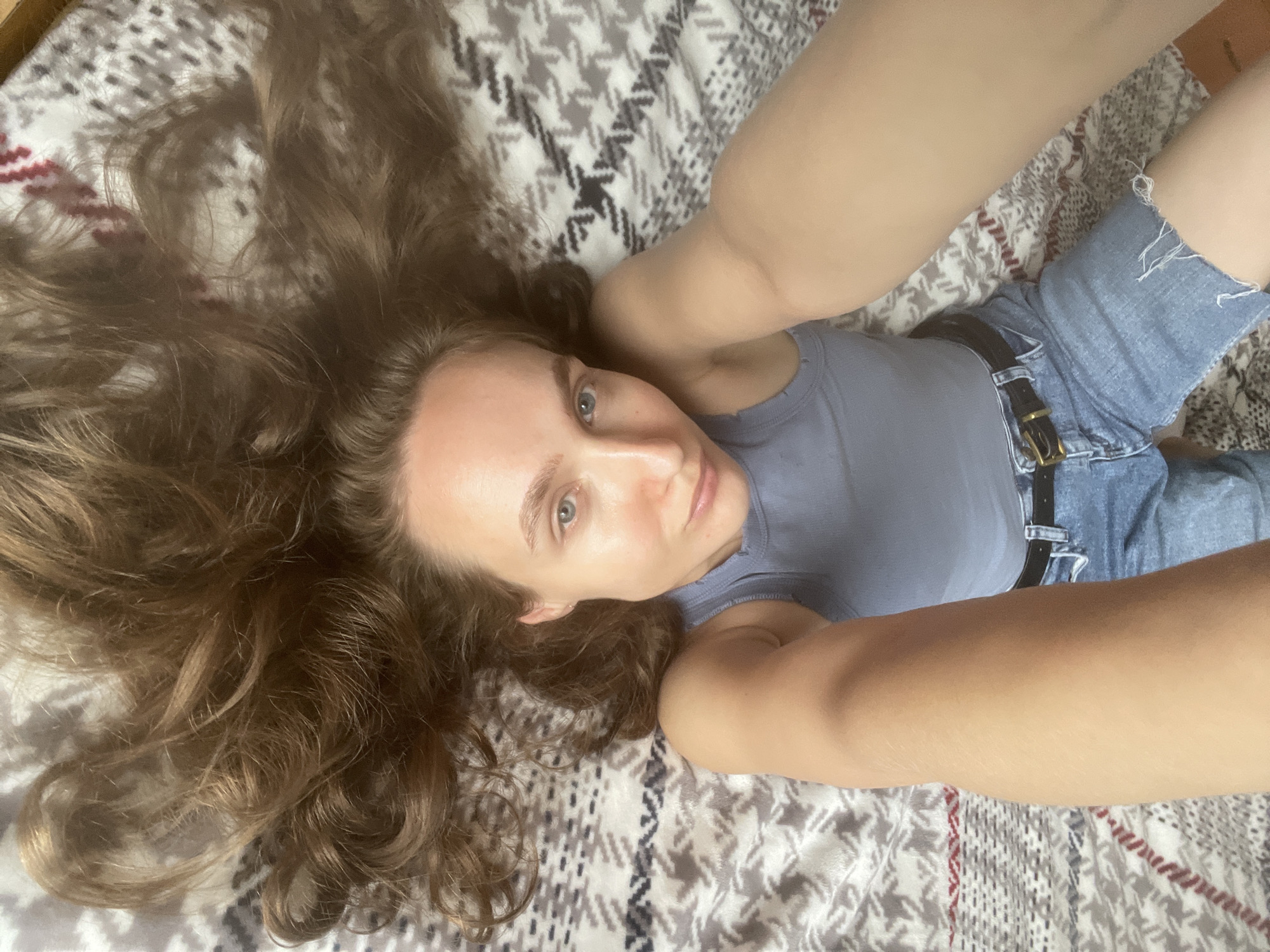 In this close-up, overhead shot, a young woman with long, wavy brown hair is lying on a patterned blanket. Her hair fans out around her head and shoulders. She is looking up towards the camera with a neutral expression. Her head is tilted slightly to her left, and her eyes are open and directed forward. She is wearing a light blue tank top and denim shorts with a black belt. Her arms are bent and extended upwards, with her hands out of view. The blanket beneath her features a geometric pattern of grey, white, and black shapes, with thin red stripes along the edges. The lighting suggests it is daytime. The setting appears to be indoors, likely a bedroom or living area. There is no visible text in the image.