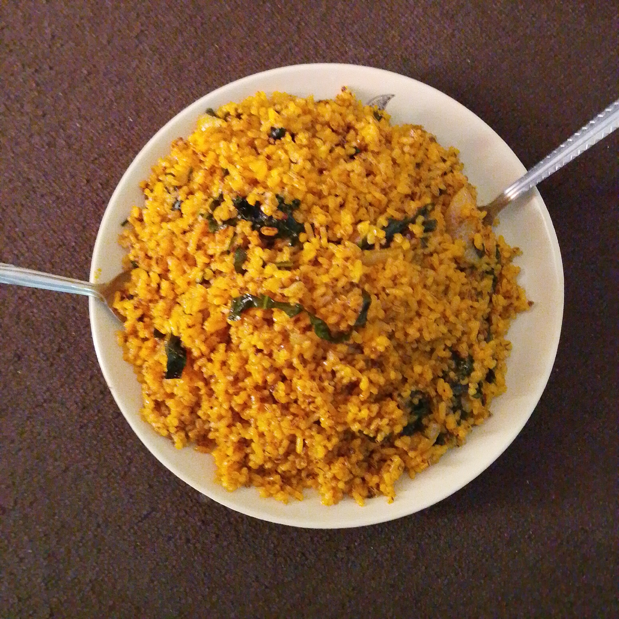 The image shows a close-up, overhead view of a white plate filled with a generous portion of yellow rice, possibly turmeric-infused, mixed with dark green leafy vegetables and some visible pieces of onion. Two metal spoons are partially visible, one on the left side of the plate, inserted into the rice, and another on the right, also in the rice. The plate is resting on a dark brown, textured surface, which appears to be a table or placemat. The lighting is somewhat diffused, casting minimal shadows. There are no people visible in the frame. The scene is likely a domestic or casual dining setting, potentially in Jalingo, Nigeria, given the context.