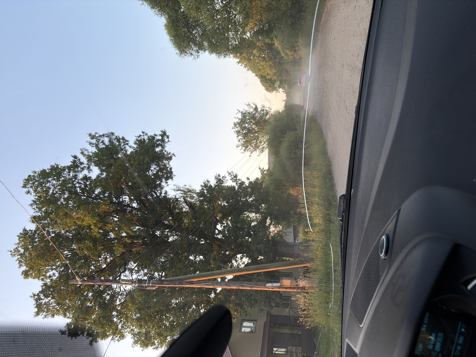 The image captures a view from inside a vehicle, looking out through the windshield. The scene depicts a rural road in the city of Nelyudovo, Russia. The road appears to be unpaved and dusty, with a single car visible in the distance creating a dust trail. On the left side of the road, there is a house with a porch and a large tree next to a utility pole. The sky is clear and light blue, suggesting it is daytime. The foreground shows the car's dashboard.