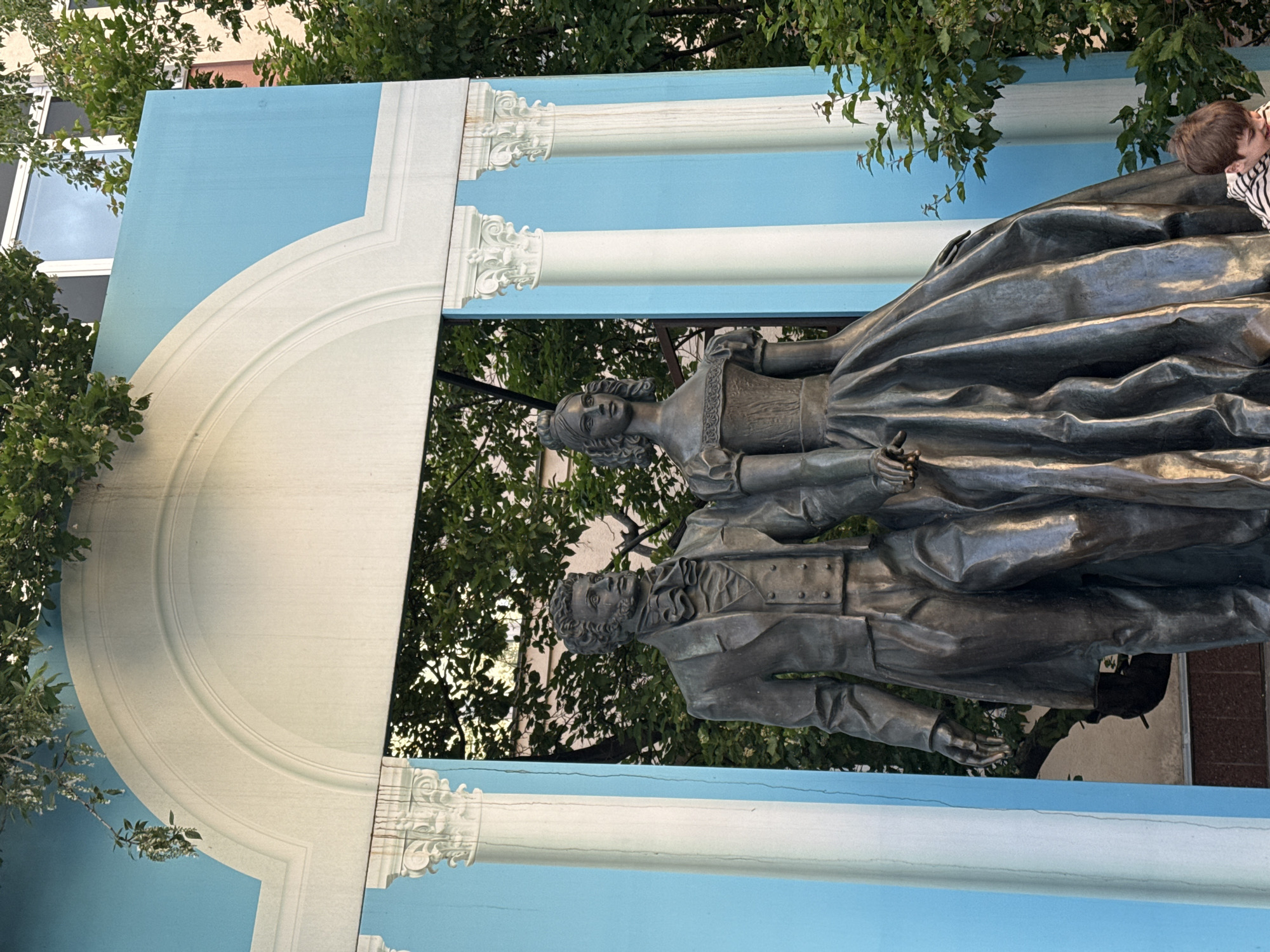 The original image is tilted on its side, presenting a bronze statue of two historical figures—a man and a woman—standing upright in front of a decorative architectural facade. The man, depicted with a beard and wearing a 19th-century coat and waistcoat, extends his left hand forward with an open palm. Beside him, the woman is adorned in a period dress with a high neckline, ruffled sleeves, and a full skirt; her hair is styled elegantly, and she holds a small item in her right hand.
The backdrop consists of a light blue wall embellished with prominent white classical architectural elements, including decorative columns with ornate capitals and a large curved archway. This painted facade creates a striking contrast with the dark, metallic texture of the bronze statue. Surrounding the monument, lush green foliage and tree branches are visible, indicating an outdoor setting, likely a public square or park. A small child, wearing a striped shirt, is partially visible on the far right, seemingly observing the statue. The scene is brightly lit, suggesting it was captured during daytime. No discernible text is visible in the image. The monument's style and setting are characteristic of historical public art found in cities like Moscow, Russia.