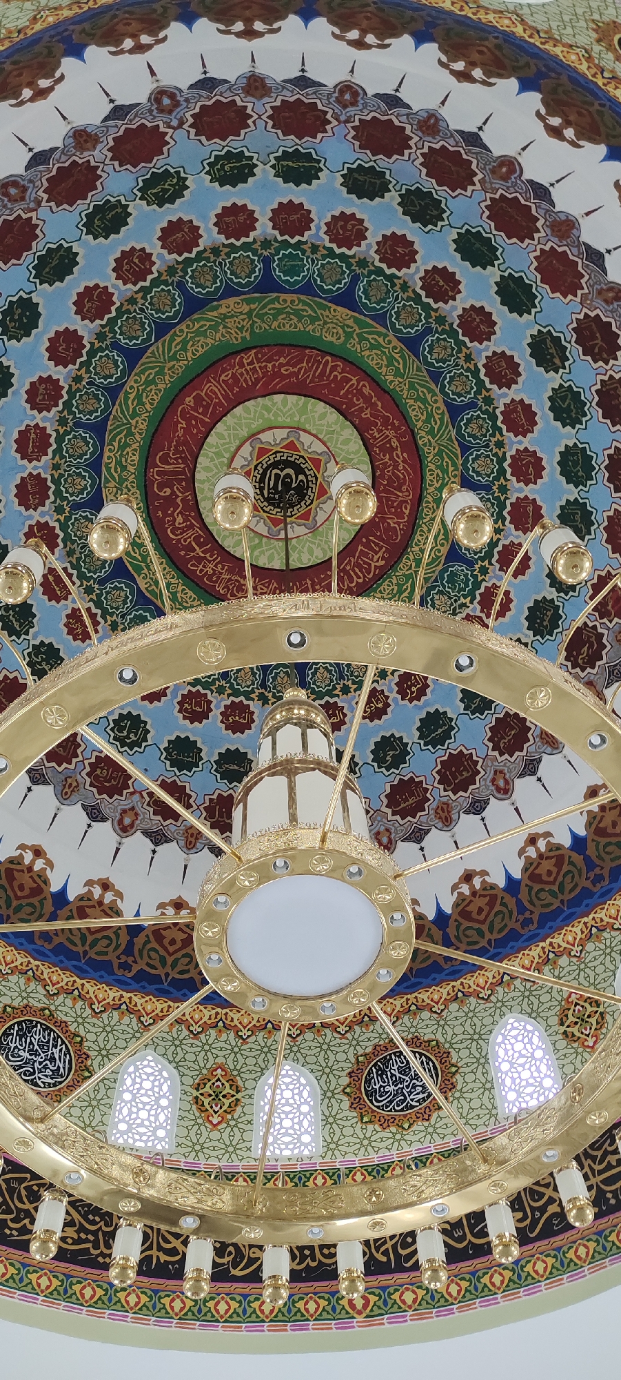 The image shows the ornate ceiling of a structure in Meunasahaluedrienlhoksukon Barat, Indonesia. The ceiling is painted with concentric circles of intricate patterns and Arabic calligraphy in red, green, blue, and gold. A large, gold-colored chandelier hangs from the ceiling. The chandelier consists of a central light fixture surrounded by a ring with smaller lights. There are also decorative window-shaped openings around the ring. The chandelier partially obscures the ceiling design.